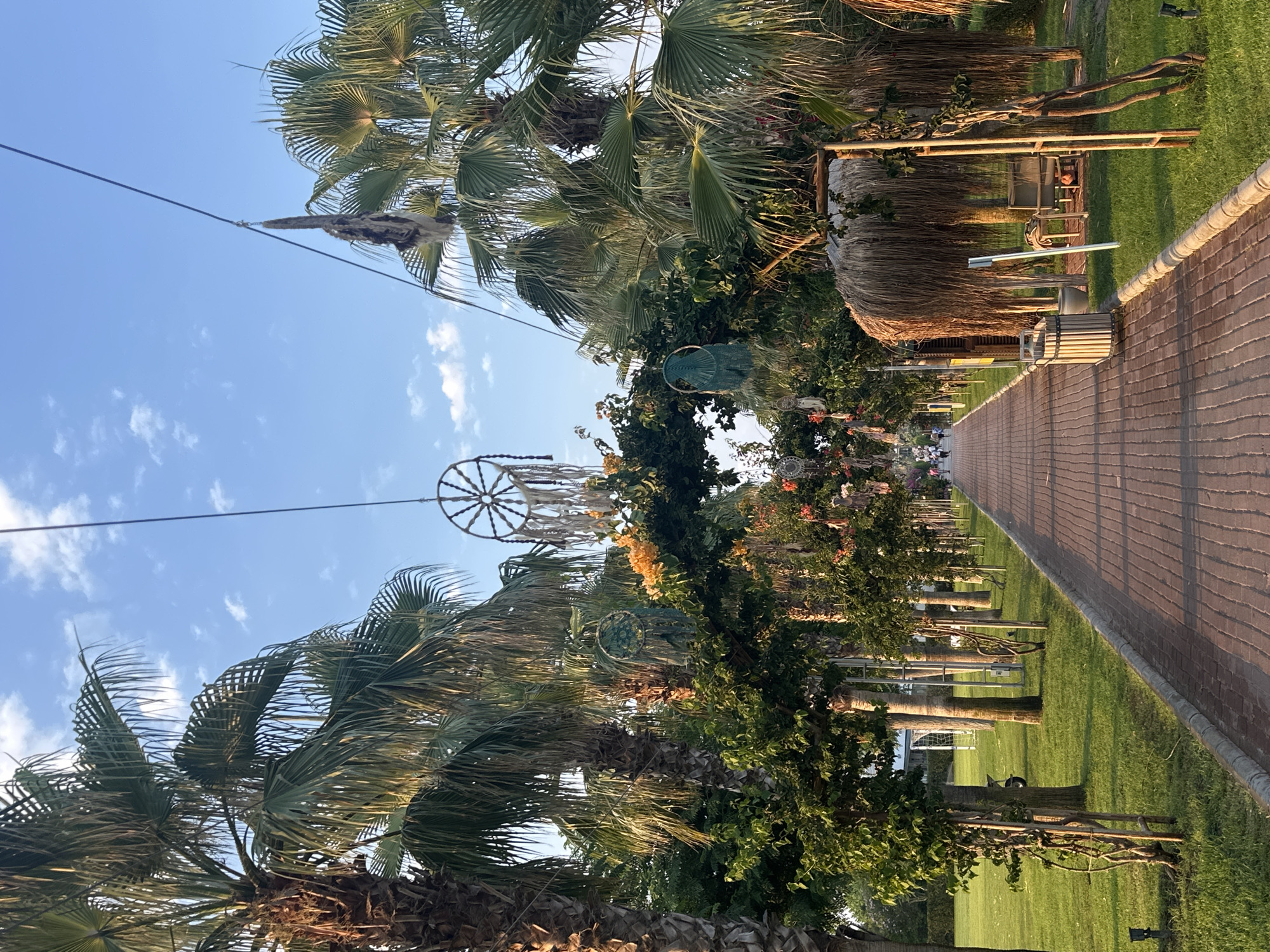 This media file depicts an outdoor scene in Manavgat, Turkey, likely during a pleasant time of day, indicated by the warm sunlight casting long shadows and the clear blue sky with scattered clouds. The setting appears to be a landscaped area with lush green grass, a paved walkway, and numerous palm trees, suggesting a resort or park environment.
The walkway, made of brick-like pavers, runs along the right side of the frame, bordered by a neatly mowed lawn. Along the left side, a line of palm trees with their characteristic fronds creates a sense of tropical ambiance. Interspersed with the palm trees and vegetation are decorative elements, including what appear to be dreamcatchers or similar macrame hangings suspended from wires or branches. These decorations, along with some floral arrangements visible in the mid-ground, suggest an event or a specially decorated area.
In the background, a thatched-roof structure, possibly a cabana or a small building, is visible, further reinforcing the resort-like atmosphere. A cylindrical trash receptacle is also positioned near this structure. The overall mood is peaceful and inviting, with a focus on natural elements and decorative touches. No specific activities are clearly depicted, but the presence of people in the distance, seated and strolling along the walkway, indicates a tranquil gathering or a leisurely day. No visible text is present in the image.