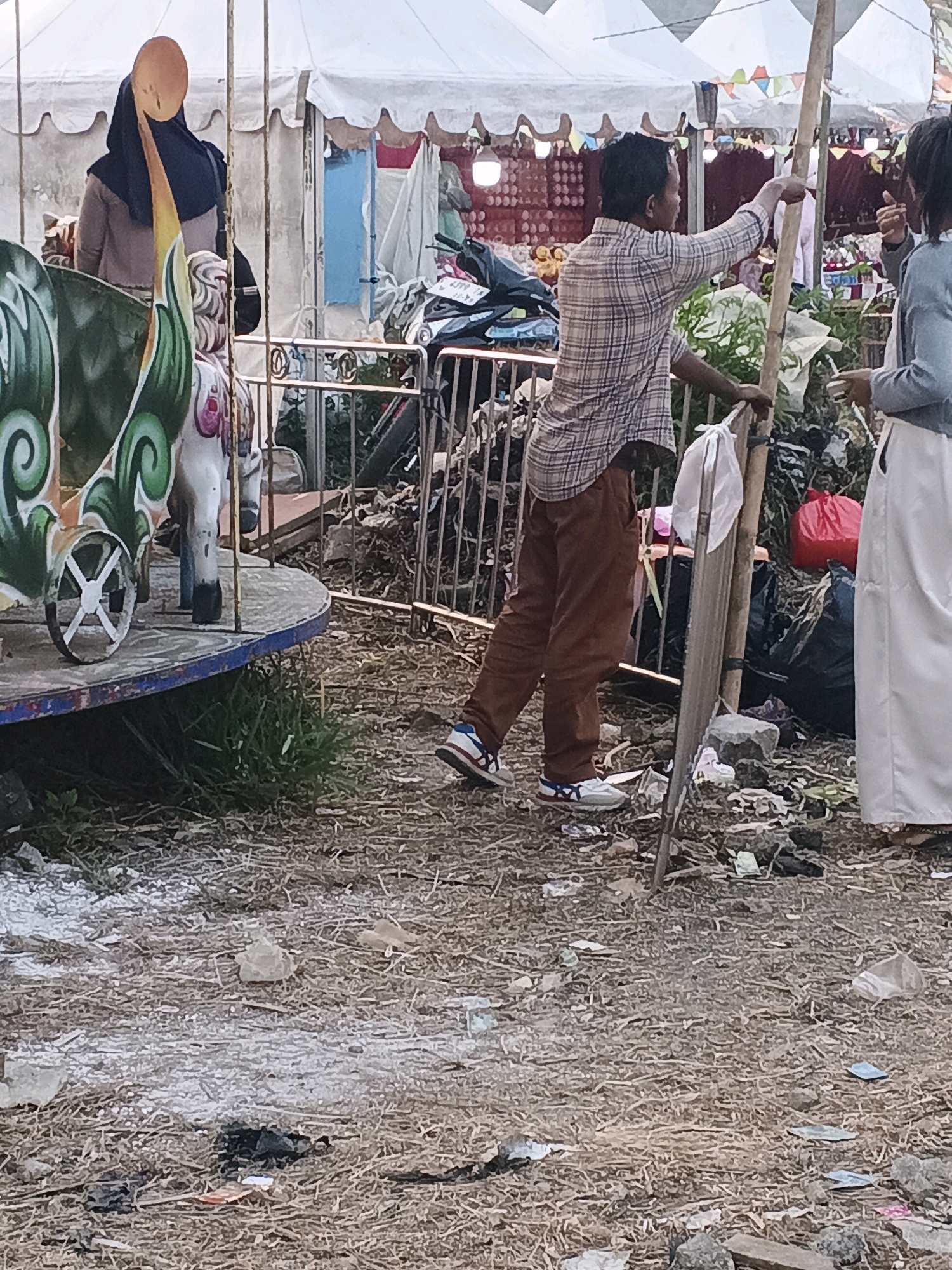 In the city of Sukabumi, Indonesia, a street scene unfolds with a carousel in the foreground. A woman, partially obscured, wears a hijab and a modest outfit. To her right, a man in a plaid shirt and brown pants stands near a makeshift stall, gesturing with his hand. Behind him, a person in a light-colored skirt stands, facing away. The ground is littered with debris, and a red plastic bag is visible. Tents, likely part of a market or fair, form the backdrop.