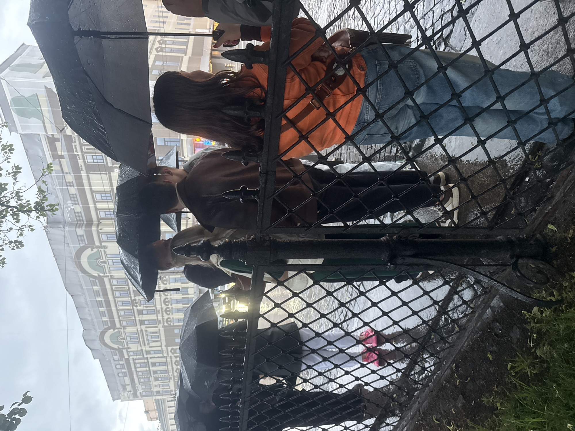 A group of people are standing under umbrellas on a wet cobblestone street in front of a black metal fence. In the background, a large, ornate building with multiple windows and arched upper stories is visible. The sky is overcast, and it appears to be raining. The people are dressed in casual clothing, and some are holding cell phones. One person in the foreground is wearing an orange jacket and blue jeans, while another is wearing white pants and bright pink shoes. The overall atmosphere is somber, likely due to the weather conditions. The building in the background, with its distinctive architectural style, suggests a location in the historic center of Saint Petersburg, Russia. The street is wet, indicating recent or ongoing rainfall, and the cobblestone pavement is typical of older European cities. The fence in the foreground is made of black metal with diamond-shaped openings. The presence of umbrellas and the wet ground are the most evident indicators of the weather.