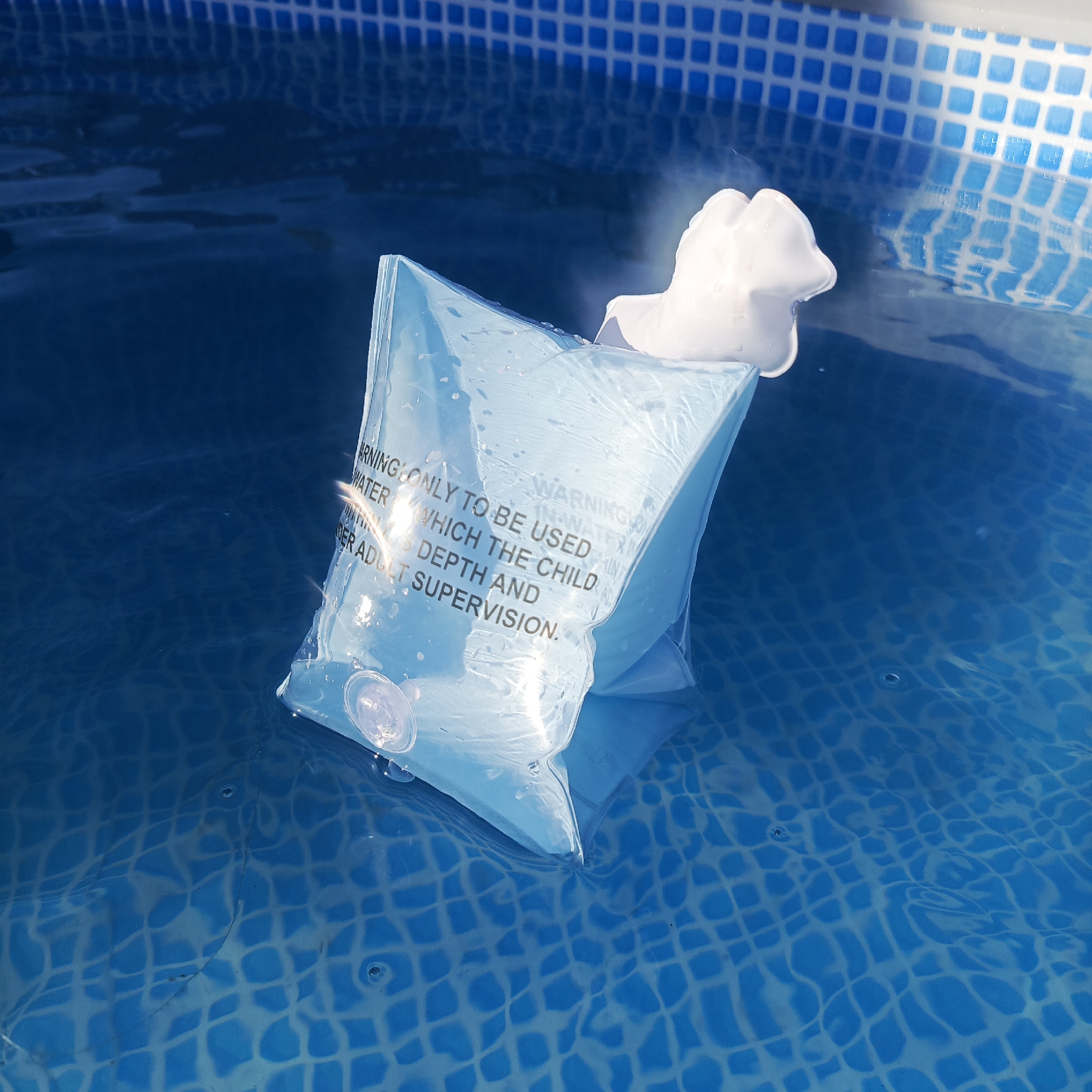 A light blue inflatable arm float with a white attachment floats in a blue swimming pool with blue and white tiled sides. The float has a warning message printed on it in black lettering, which reads: "WARNING ONLY TO BE USED IN WATER WHICH THE CHILD DEPTH AND DER ADULT SUPERVISION."