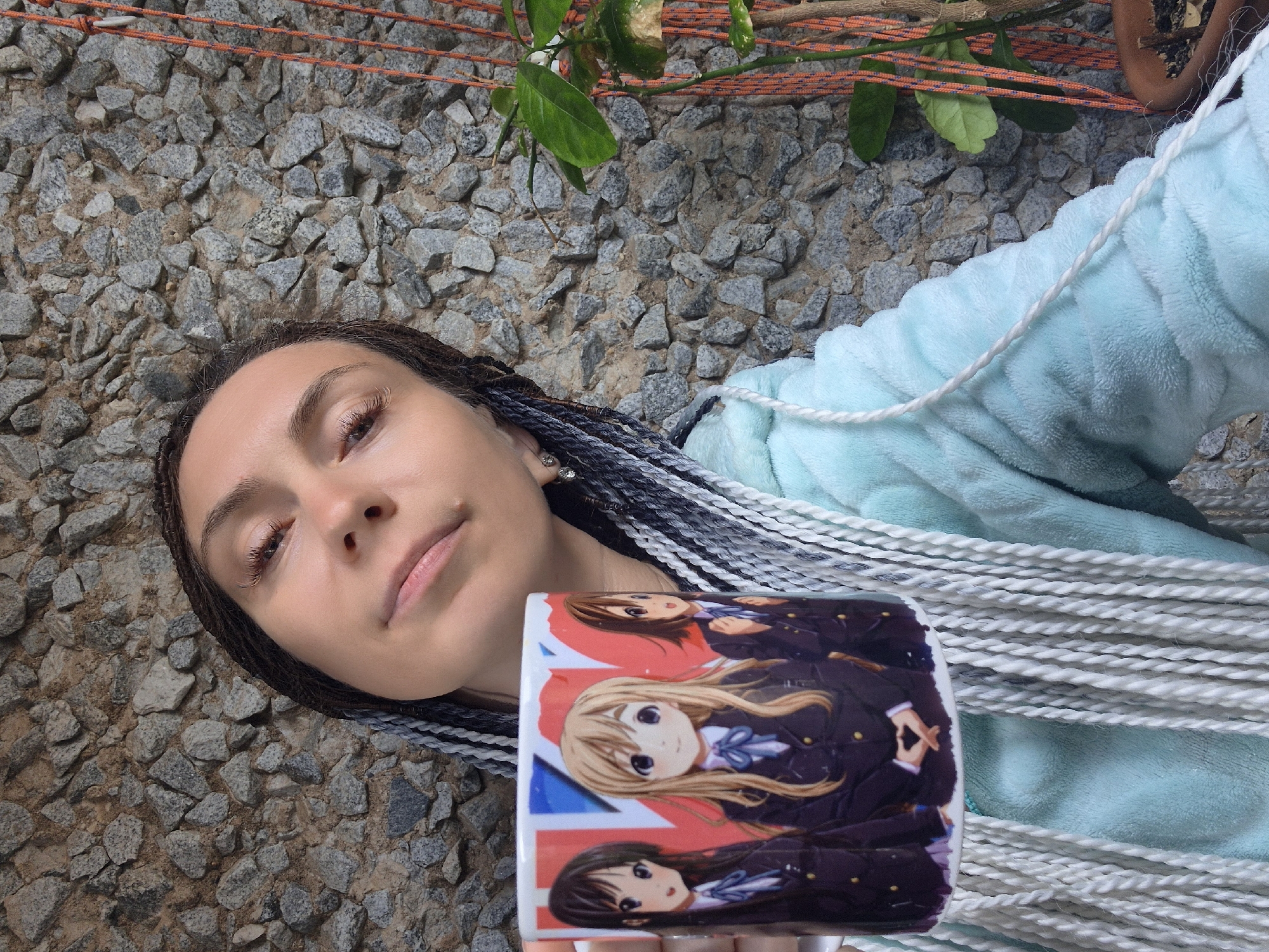 A person with braided hair is holding a mug with anime characters on it. The background is a stone wall with some plants and orange ropes. The person is wearing a light blue robe. The image appears to be a selfie. The location is Chaikovsky, Russia.