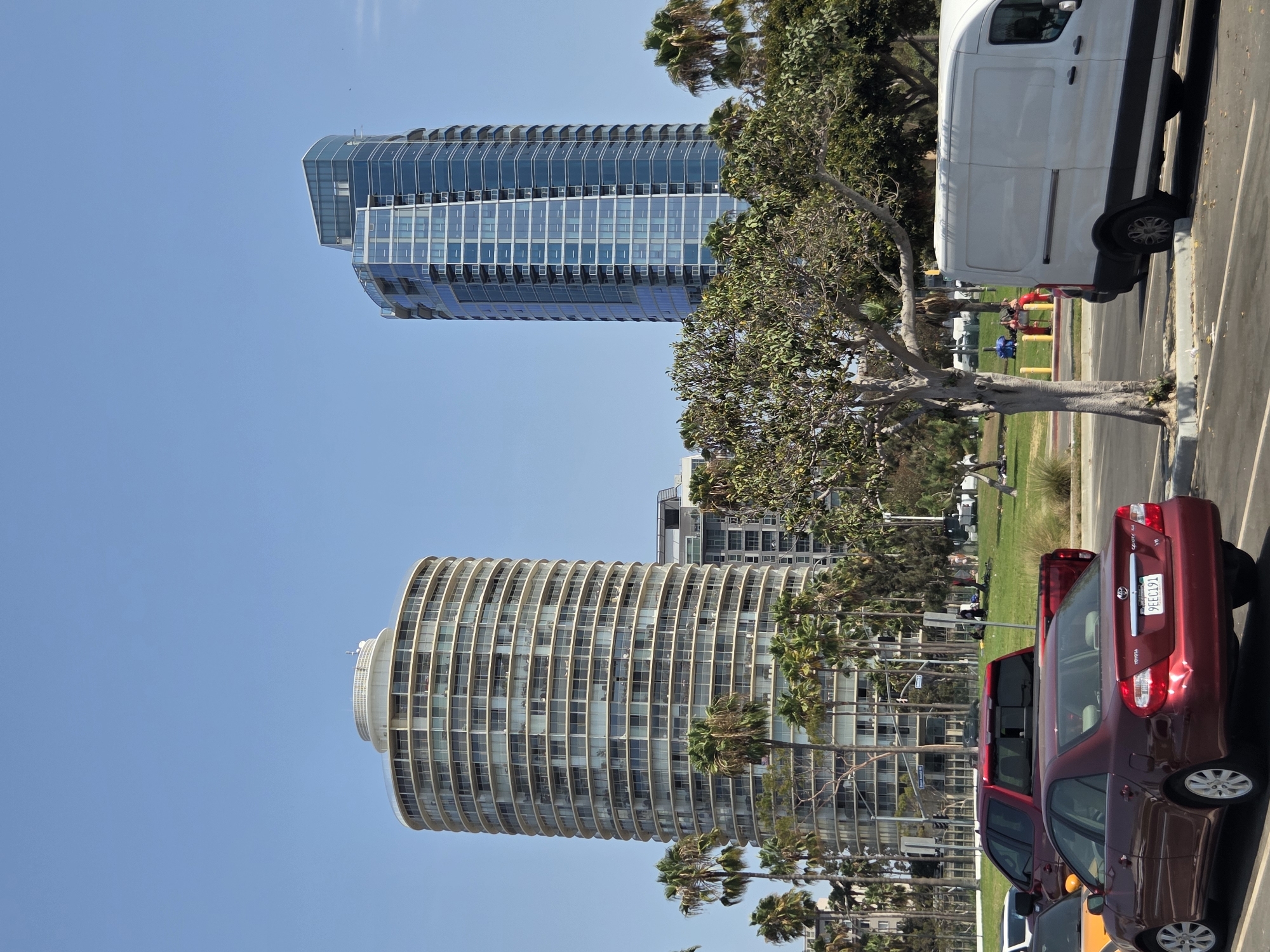 The image captures a sunny daytime scene in Long Beach, California, featuring a park and high-rise buildings. In the foreground, several cars are parked and driving on a paved surface. A red Toyota Camry with license plate 9EEC191 is prominently displayed, along with a red pickup truck. Further back, a white van is parked. The middle ground shows a grassy park area with palm trees and other foliage. In the background, two distinct skyscrapers dominate the skyline. On the left, a circular, light-colored building with numerous balconies is visible, and to its right, a taller, modern blue glass skyscraper stands. The sky is clear and blue.