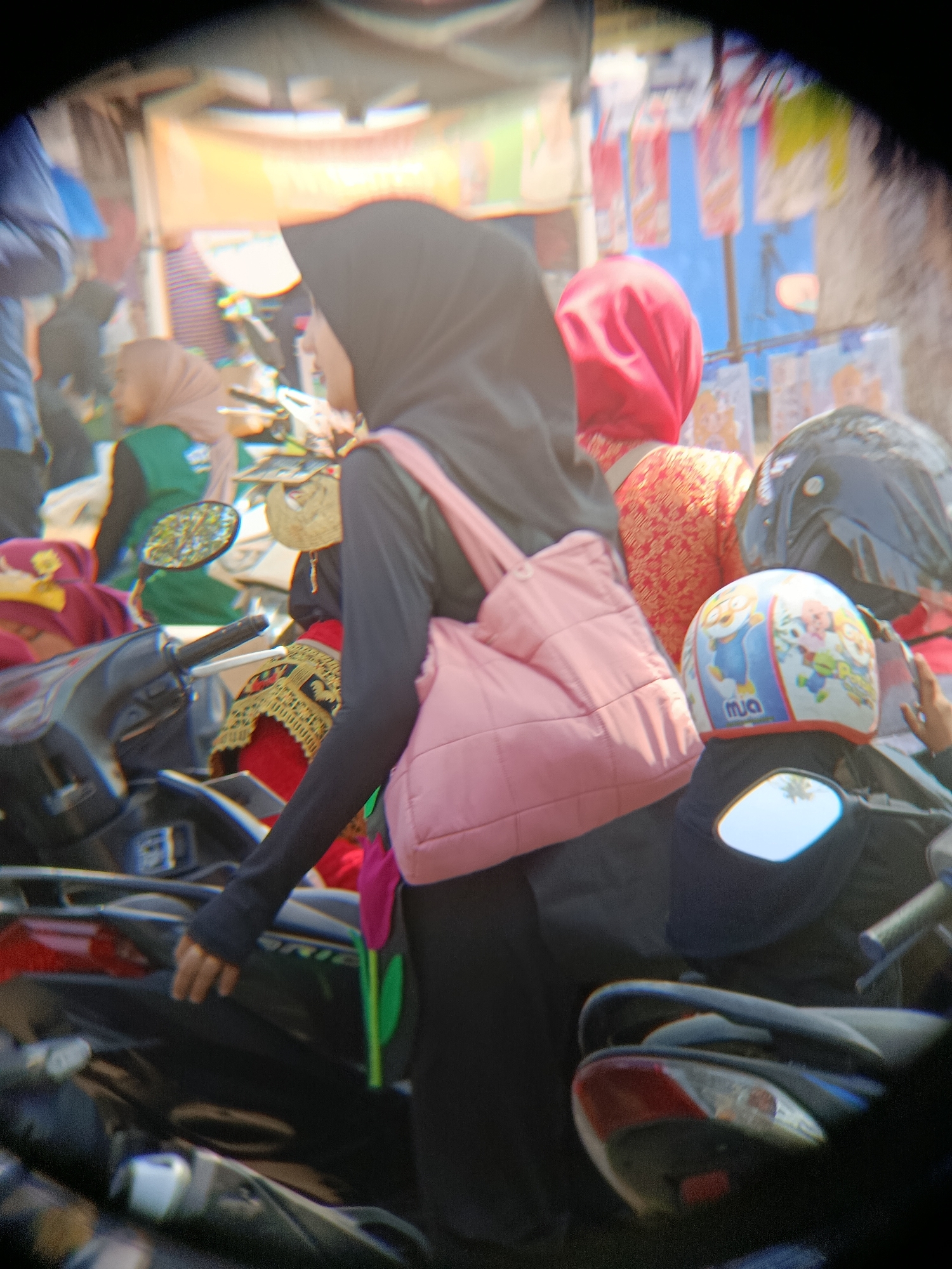 In Desa Kebon Manis, Kecamatan Cilacap Utara, Indonesia, a group of people are gathered outdoors, with several motorcycles visible. The focus is on a woman wearing a black hijab and a pink quilted bag slung over her shoulder. She is standing next to a motorcycle, and her hand is resting on the handlebars. To her right, another person wears a red hijab and a patterned orange top, and behind them, a black motorcycle helmet and another helmet with cartoon characters are visible. The background suggests a bustling outdoor market or gathering area, with banners and stalls. The lighting is bright, indicating daytime.