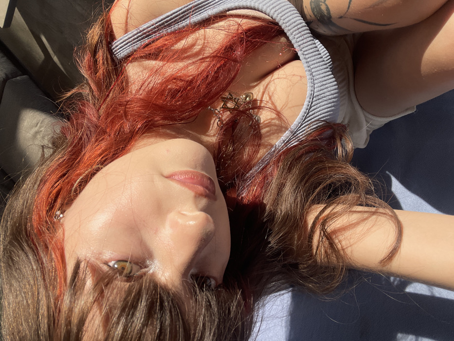 A close-up, high-angle photograph depicts a female individual with medium skin tone, partially visible from the chest up, reclining indoors in Raanana, Israel. Her long, wavy hair is predominantly dark brown with significant red/orange highlights, particularly on the top and sides, with brown bangs partially obscuring her forehead and left eye. Her left eye, featuring a brown-green iris, is open and directed towards the camera. Her lips are glossy and light pinkish-brown. A small metallic earring is visible in her upper left ear. She wears a light gray ribbed tank top, revealing cleavage, and a metallic necklace with multiple charms, including a Star of David and a small bell-shaped object, is visible in her décolletage. An indistinct dark tattoo is visible on her right upper arm/shoulder. Her left forearm rests on a deep blue fabric surface in the lower left of the frame. Strong, directional sunlight illuminates her left side, creating bright highlights and casting distinct shadows. A dark gray upholstered object is visible in the upper right background.