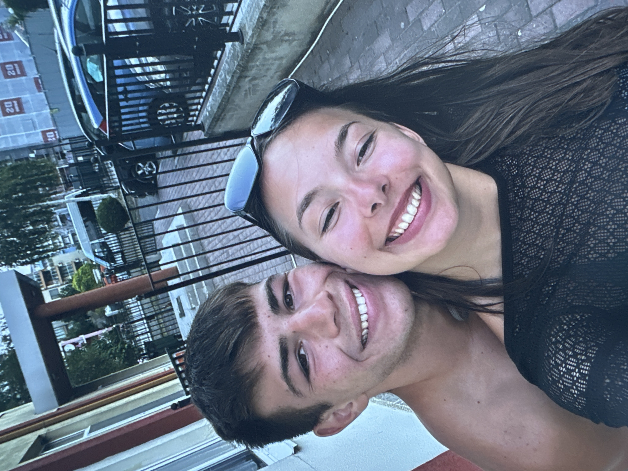 A close-up, waist-up, outdoor, selfie-style photograph features a young Caucasian couple standing close together, with the woman slightly in front of the man. Both are smiling broadly at the camera. The man, on the left, has short, dark brown hair and is shirtless, revealing his shoulders and upper back. He has a light tan with some freckles. The woman, on the right, has long, dark brown hair that falls over her shoulders. She is wearing a black, sheer, lace-like top. A pair of black sunglasses are perched on top of her head.
The background suggests an urban setting. To the left, a building facade is visible with red and yellow trim. Behind them, a black metal fence encloses an area with paving stones. Further in the background, a blue car is parked, and other vehicles and buildings can be seen, some with construction scaffolding and signage. The overall lighting suggests it is daytime, possibly late afternoon or early evening. The photograph appears to be taken on a street or walkway, with the paving stones of the walkway visible in the foreground and to the right.
The couple is likely on a street in Sofia, Bulgaria, given the context provided.