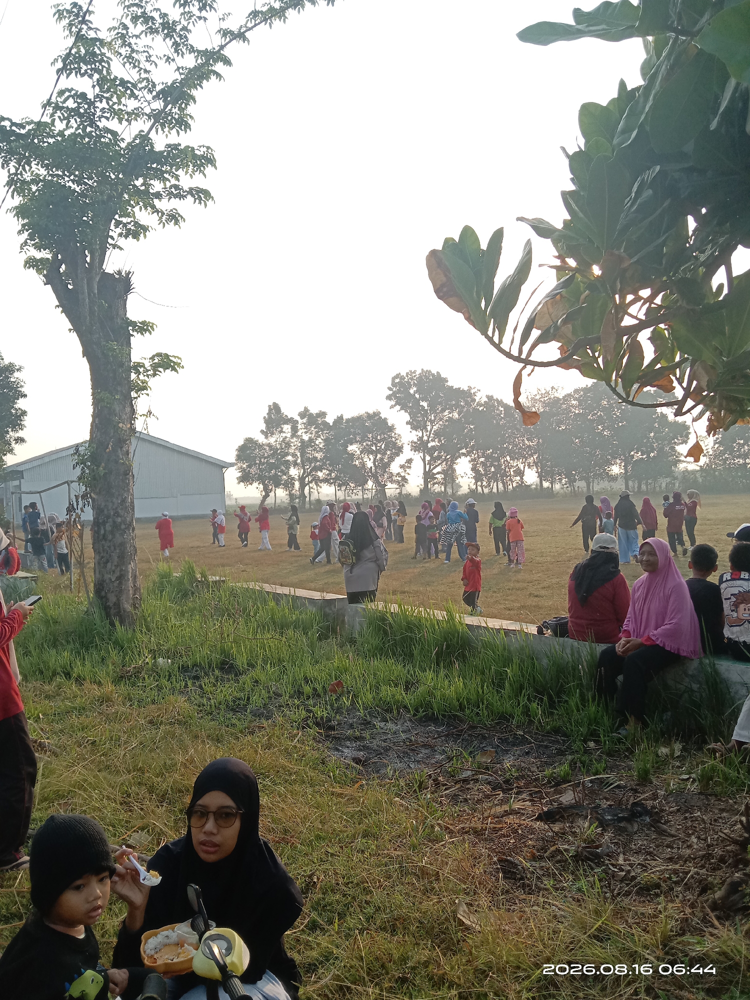 In a grassy field with trees in the background, a group of people, including children and adults, are gathered outdoors. Some individuals are seen standing and engaging in activities in the middle ground, while others are seated in the foreground. Two people in the immediate foreground are eating from a container. The scene appears to be illuminated by natural daylight, possibly early morning given the soft light and potential mist. The timestamp on the image indicates "2026.08.16 06:44", and the location is identified as Japanan, Indonesia.
