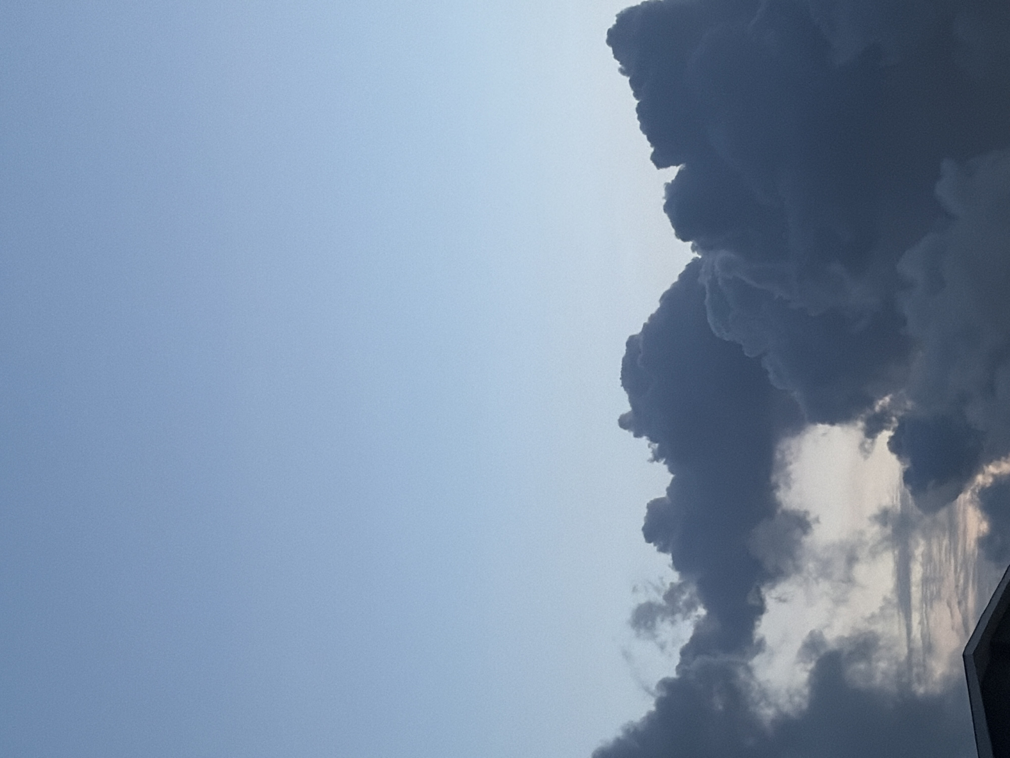 The image shows a sky with a large, dark cumulonimbus cloud formation dominating the right side. The sky is mostly pale blue on the left. The bottom right corner includes part of a structure with a straight edge. The image was taken in Igboefon, Nigeria. There are no people visible.