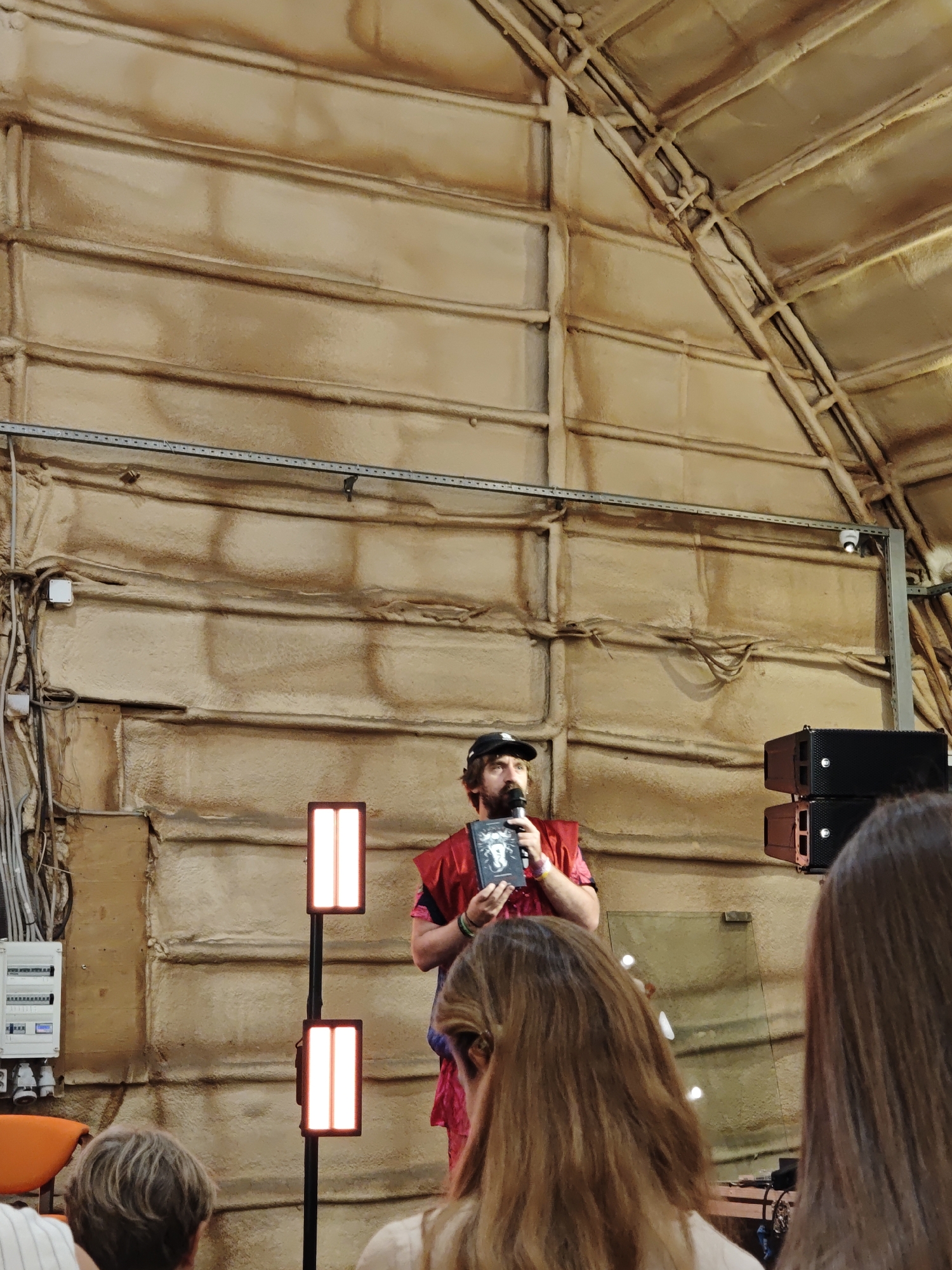 A man wearing a black baseball cap and a red outfit is standing on a stage, holding a book and speaking into a microphone. He has a beard and appears to be in his 30s or 40s. Behind him, the wall is textured and appears to be made of a beige material. There are two tall, rectangular lights with illuminated panels on the left side of the stage. On the right side, there are two black speakers stacked on top of each other. In the foreground, the backs of several audience members' heads are visible. The overall environment suggests an indoor event or performance space. The location is not specified in the image.