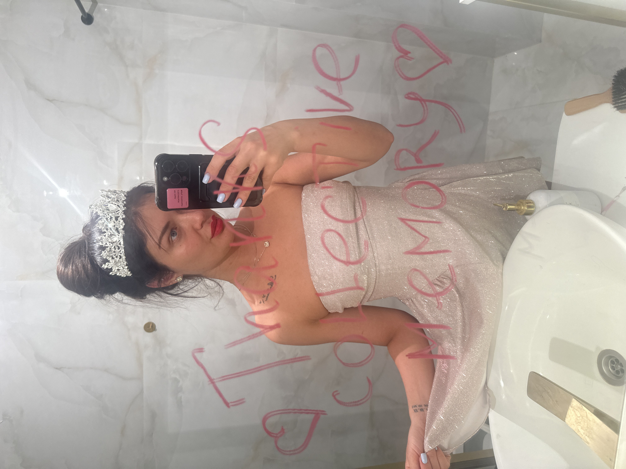 A young woman, dressed in a sparkling, strapless light-colored gown and a silver tiara, takes a mirror selfie in a bathroom. Her dark hair is styled in an updo, and she wears red lipstick and light blue nail polish. She holds a black smartphone with multiple camera lenses to capture her reflection. A delicate necklace and a small tattoo on her left wrist are visible.
The background reveals a bathroom interior with white tiled walls featuring a subtle marble-like pattern. A white porcelain sink with a gold-toned faucet and drain is seen on the right. On the sink's counter, there's a small white bottle with a gold pump dispenser and a hairbrush with a wooden handle. A black pipe or fixture is visible in the upper left corner.
Written in pink, backward (as seen in the reflection), on the mirror is the text: "I LOVE COLLECTIVE HARMONY <3". The woman maintains a neutral expression, looking directly into the camera. The scene suggests an indoor setting, possibly in Khimki, Russia, and the attire implies preparation for a special event or celebration.