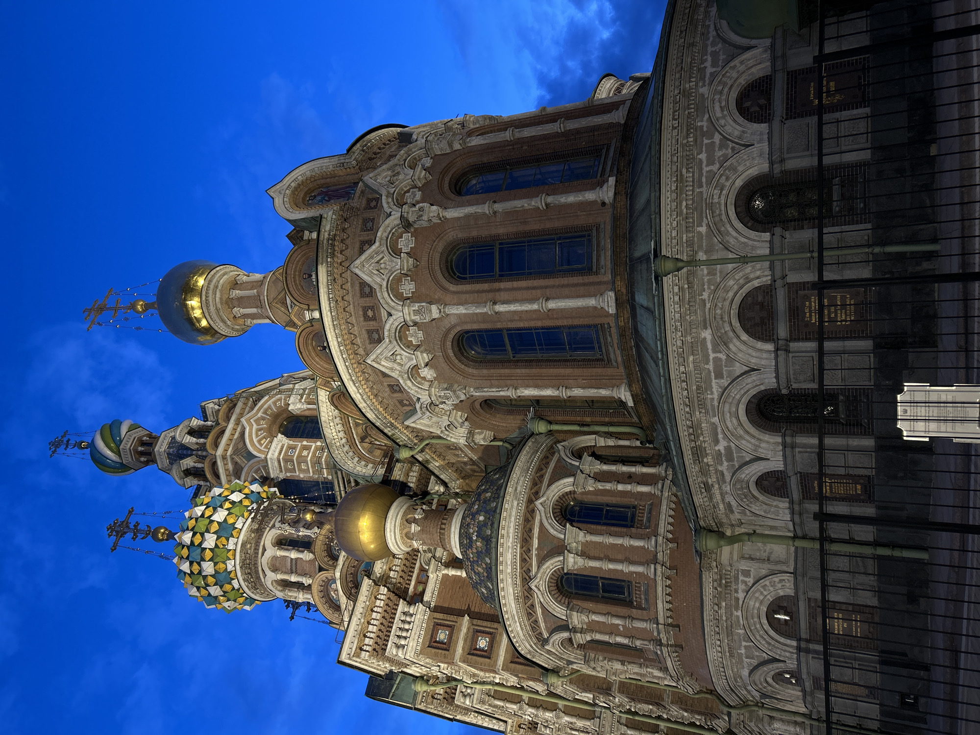 The media file presents an exterior, low-angle photograph of a grand, ornate Russian Orthodox church, identified as likely the Church of the Savior on Spilled Blood in Saint Petersburg, Russia. The image captures the building at twilight or early night, with the sky a deep, rich blue featuring faint cloud formations, creating a dramatic contrast with the illuminated structure.
The church's distinctive Russian Revival architecture is prominent, characterized by multiple onion domes. Several domes are visible, including one with a brilliant golden finish and others adorned with intricate mosaic patterns of blue, green, and white tiles. Each dome is crowned with an ornate Orthodox cross. The facade below is extensively decorated with a mix of red brickwork, white stone carvings, arches, columns, and detailed mosaic panels, likely depicting religious iconography. Numerous arched windows, some dark and others subtly catching light, are integrated into the elaborate design.
In the foreground on the right, a dark metal fence or railing runs horizontally, partially obscuring the lower section of the building. A traditional street lamp with a green pole is also visible near the fence. There are no people present in the scene, which focuses entirely on the architectural splendor.
The lighting conditions highlight the intricate details and textures of the church's decorative elements against the darkening sky, emphasizing its historical and artistic significance. Behind the fence, several dark plaques or signs are visible on the building's lower wall. While most Cyrillic text on these plaques is indecipherable due to distance and resolution, one prominent set of dates, "1970 - 1980", can be clearly read.