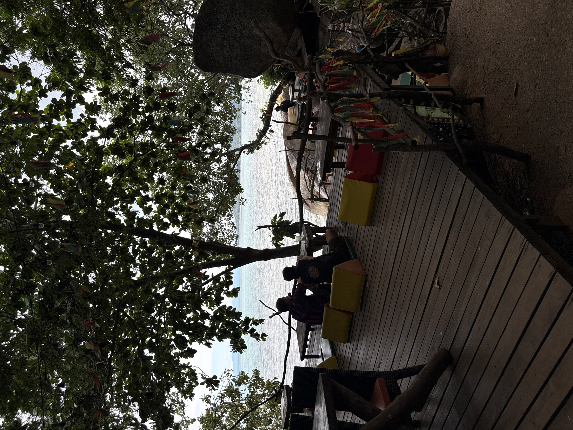 The image depicts a rustic, open-air dining or seating area next to a body of water. A wooden deck forms the primary structure, with several tables and seating arrangements. Some of the seating consists of brightly colored cushions, yellow and red, placed on low platforms or benches. Decorative elements, such as colorful fabric strips, are strung along railings and branches of nearby trees, suggesting a festive or tropical atmosphere.
In the foreground, a large tree with dense foliage partially obscures the view, creating dappled light and shadow. The water in the background appears calm and reflects the bright sky, indicating fair weather. The scene suggests a daytime setting, possibly late morning or early afternoon given the brightness.
Several people are visible, seated at tables on the deck, engaged in conversation or relaxation. They are silhouetted against the bright background, making their features indistinct. One group appears to be seated together, while others are more spread out. The overall mood seems relaxed and casual, consistent with a beachfront or waterside establishment in a tropical location like Baan Hua Thanon, Thailand. The presence of a large boulder near the seating area further anchors the location to a natural, perhaps rocky, coastal environment. The image is oriented sideways.
