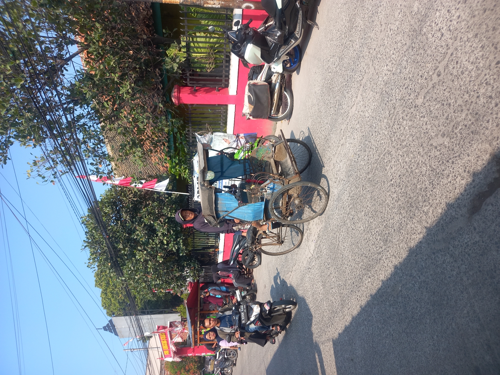 In Linggar, Indonesia, a busy street scene unfolds under a clear blue sky. A man in a dark hoodie is driving a three-wheeled pedicab, its blue canopy shielding him from the sun. He wears a light-colored cap and appears to be looking straight ahead. In front of the pedicab, a motorcycle speeds by with two passengers. The man driving wears a blue shirt and jeans, while the woman behind him wears a dark hijab and a black long-sleeved shirt. Further down the street, other vehicles and people are visible, including a parked motorcycle with a white and black body and blue wheels, and a red building with a fence in the background. Indonesian flags, red and white, are displayed on poles. The road is paved with asphalt, and a shadow of a building stretches across the surface, indicating the time of day. The overall impression is one of daily life in a bustling Indonesian city.
