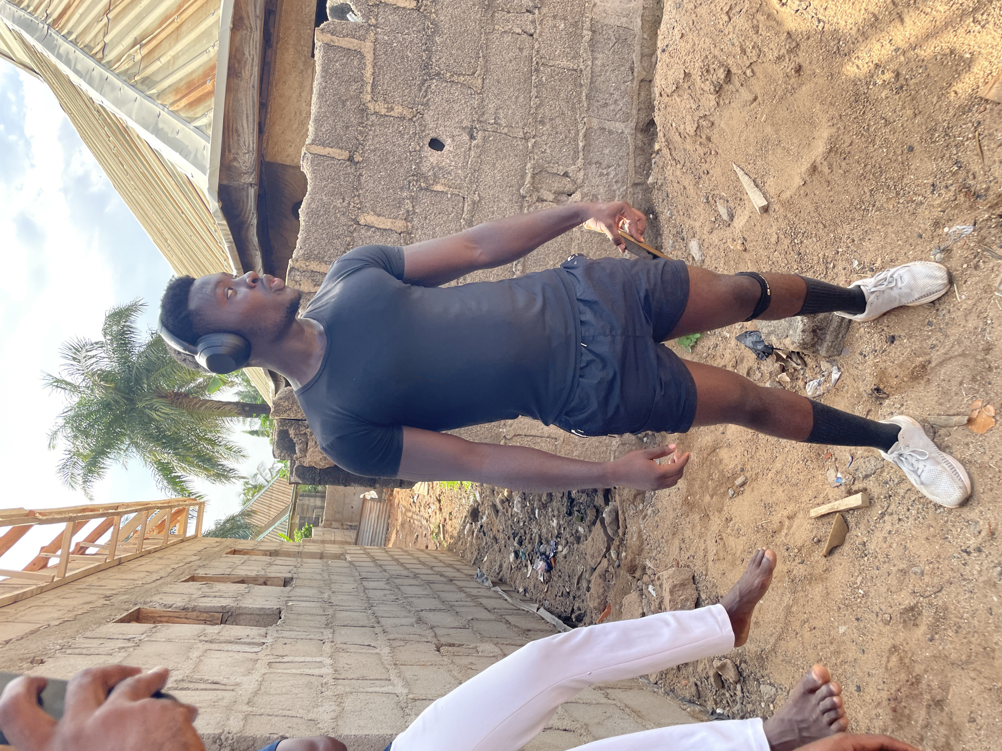 A medium shot captures a man wearing a black t-shirt, black shorts, black socks, and white shoes. He is also wearing headphones. He is standing on a sandy surface that includes bits of trash and stones. Beside the man, there are two bare feet visible in the lower portion of the shot. A person wearing white pants is also visible in the foreground. In the background there is a grey cinder block wall, corrugated metal roofing, and a palm tree visible against the sky. The shot appears to be taken in the city of Akwanga, Nigeria.