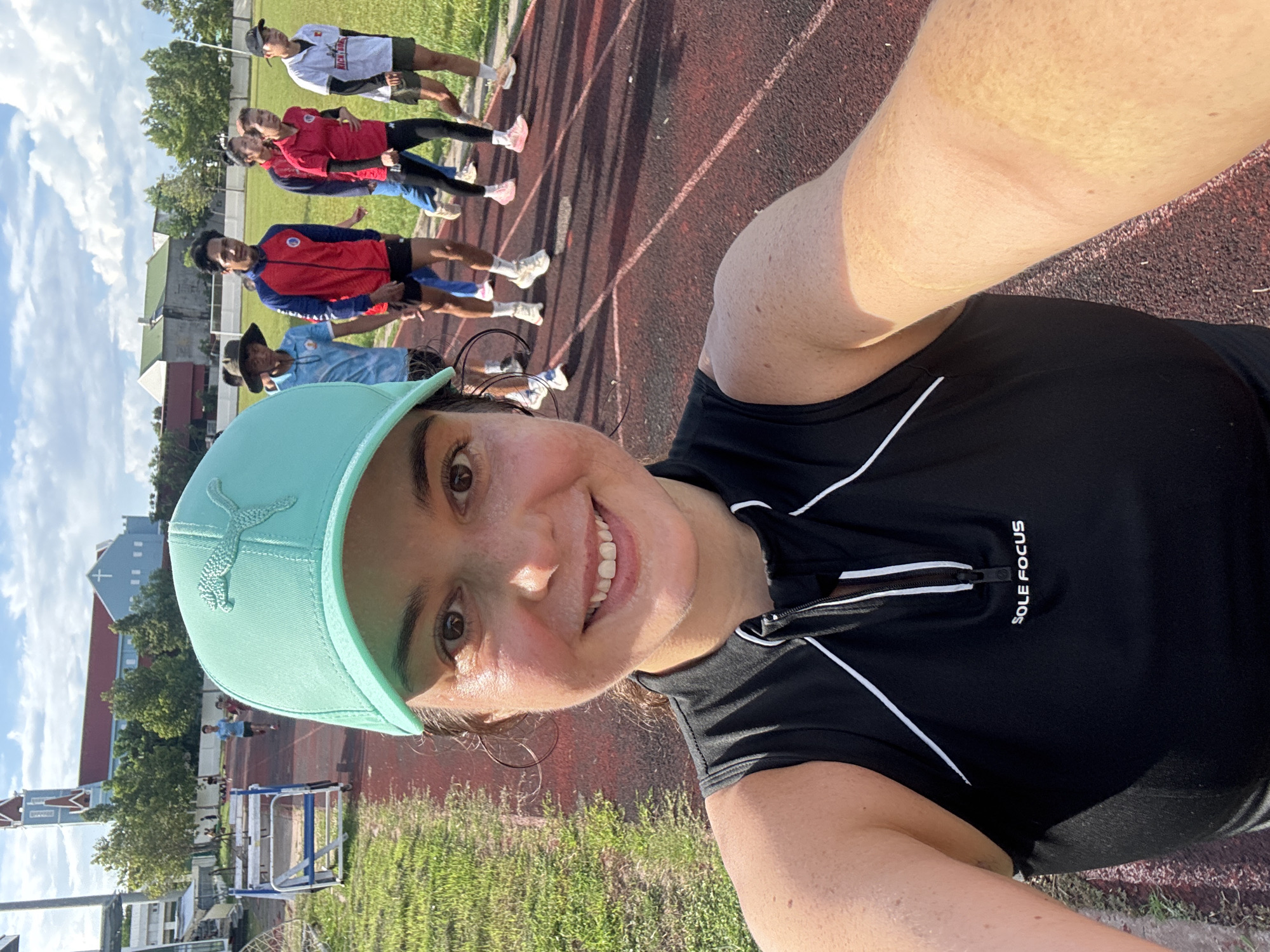 This image captures a group of individuals on a red running track in an outdoor sports stadium, likely located in Phan Thiết, Vietnam, given the presence of a distinctive blue and red-roofed church with a steeple in the background. The sun is shining, casting shadows on the track.
In the foreground, a woman wearing a teal baseball cap and a black sleeveless athletic top with white trim is taking a selfie. She has dark hair and is smiling. Behind her, a group of people are walking or running on the track. The individuals are varied in gender and ethnicity, and appear to be engaged in athletic activity. They are wearing athletic clothing, including t-shirts, jackets, shorts, and track pants, in various colors. Some individuals are wearing hats.
To the left of the track, there are hurdles set up on a grassy area. In the distance, buildings and trees are visible. The sky is partly cloudy. The overall scene suggests a training session or a recreational sports event.