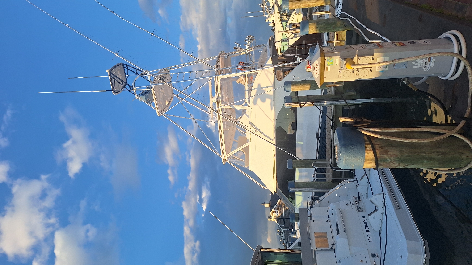 This image depicts a marina scene, likely in Nantucket, United States of America, given the context of the provided crops. The foreground features a wooden dock with several pilings, one of which is wrapped with a thick rope and a coiled hose. A utility pedestal, possibly for electrical and water hookups, is positioned on the dock, with cables connected.
In the mid-ground, two white boats are docked. The boat on the left appears to be a smaller vessel with "EAST GREENWICH, RI" visible on its stern. To its right, a larger, more prominent white yacht is moored. This yacht has a tall, multi-tiered structure with numerous support poles and rigging, indicative of a fishing or cruising vessel. The yacht's cabin is enclosed with clear plastic sheeting.
The background shows other boats and buildings, including structures with peaked roofs and a steeple, suggesting a waterfront town. The sky above is a vibrant blue, dotted with scattered white clouds, and illuminated by warm sunlight, indicating it is either early morning or late afternoon. The overall impression is a peaceful maritime setting.