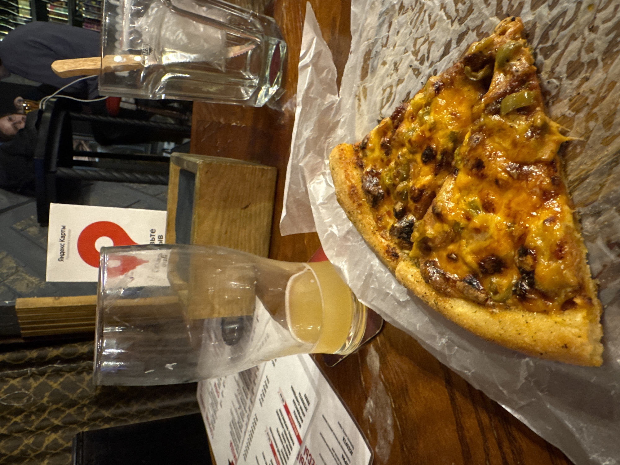 The image shows a slice of pizza, likely from the 1932 ЭСКИЗЫ restaurant located in Chaikovsky, Russia, judging by the menu visible. The pizza is on a paper liner. A glass of beer or cider sits next to the menu. Other items on the table include additional glassware, a wooden cube, and what seems to be a card with the "Яндекс Карты" logo. In the background, there are blurred figures seated at a table in the restaurant.