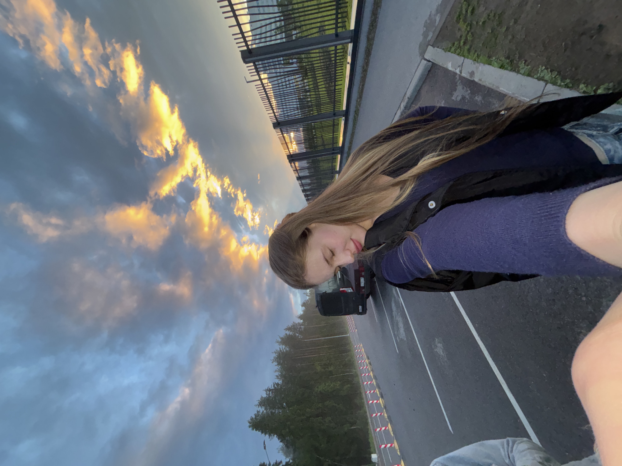 A young woman with long brown hair is pictured outdoors at dusk in a parking lot, with a striking sunset visible in the background. She is wearing a dark jacket over a blue sweater and patterned pants. The sky is filled with dramatic clouds, with vibrant oranges and yellows illuminating the scene. A black van and a red car are parked in the background, along with a fence and some buildings. The location appears to be in or near Arfa, Russia, given the context of the request.