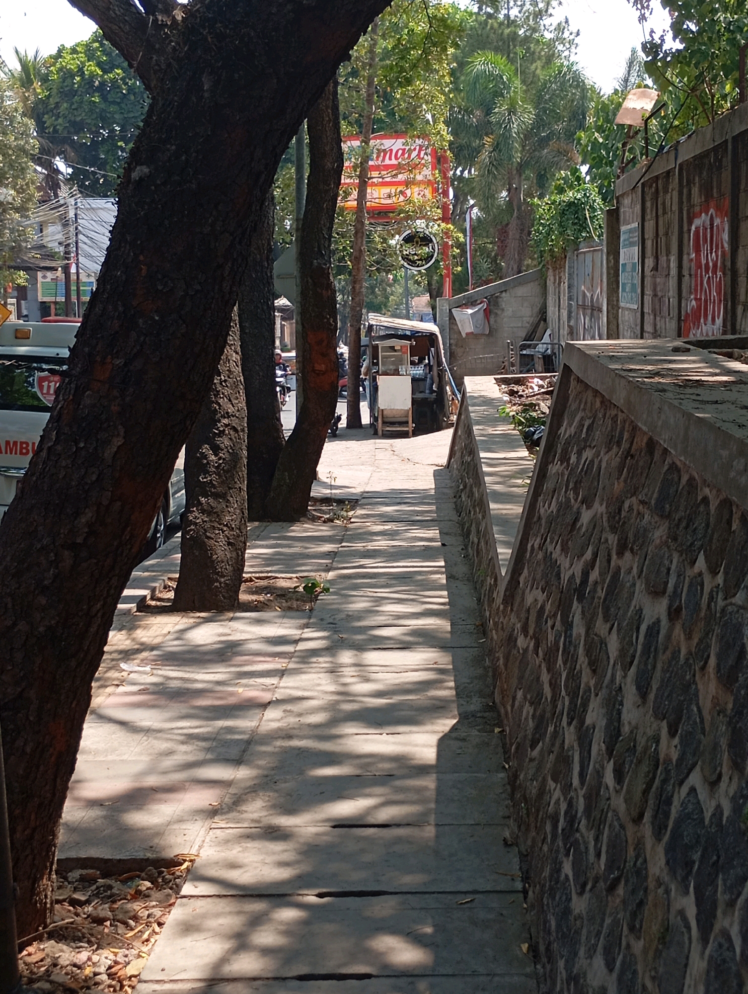 The image depicts a sidewalk in the city of Cimahi, Indonesia, lined with trees on the left and a stone wall on the right. Sunlight filters through the trees, casting shadows on the concrete sidewalk. In the background, a street scene unfolds with a small food cart, a vehicle with "AMB" visible on its side, and a motorcycle with a rider. A red sign with the word "mart" is prominent, indicating a commercial establishment. Graffiti is visible on the stone wall. The environment appears to be an urban setting, likely during the daytime.