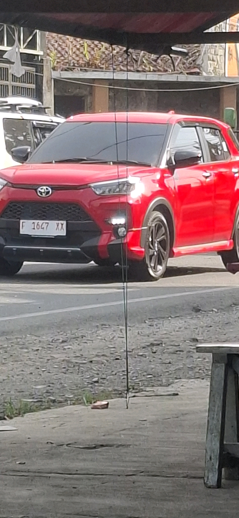 A red Toyota Raize is parked on a paved road in what appears to be an outdoor market or roadside area in Rancagoong, Indonesia. The car has a license plate that reads "F 1647 XX". The vehicle is bright red with black accents on the lower bumper and wheel arches. The headlights and fog lights are illuminated. A thin, vertical wire or string is visible hanging in front of the car. In the background, there is a building with a textured wall and some fencing. A portion of another vehicle, possibly white, is visible to the left of the red car. The ground in the foreground is a mix of pavement and gravel with some scattered debris and vegetation. A wooden structure, possibly a table or bench, is partially visible on the right side of the image.