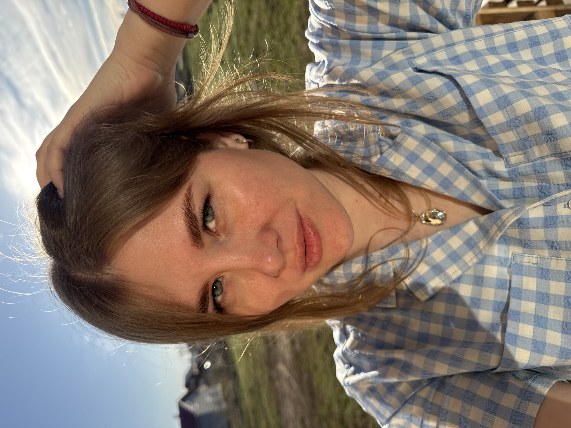 A young woman is posing for a photo in an outdoor setting in Ферма, Russia. She has fair skin, brown hair, and blue eyes. She is wearing a blue and white gingham shirt and a red bracelet on her wrist. She is holding her hair up with one hand. The background includes a cloudy sky, green grass, and some buildings.