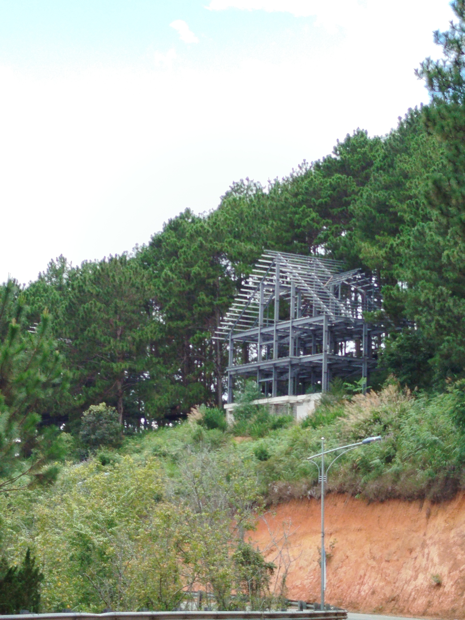 The image shows a multi-story metal building frame under construction, set against a backdrop of dense evergreen trees on a hillside. The structure appears to be a house or a small building, with exposed beams and supports. Below the construction site, a bright orange-brown dirt embankment slopes down towards a road. A modern street lamp with a curved arm stands on the roadside, its light directed towards the street. The sky is overcast and pale blue, with a few wispy white clouds. The overall scene suggests a semi-rural or developing area, potentially on the outskirts of Da Lat, Vietnam, a city known for its pine forests and hilly terrain.