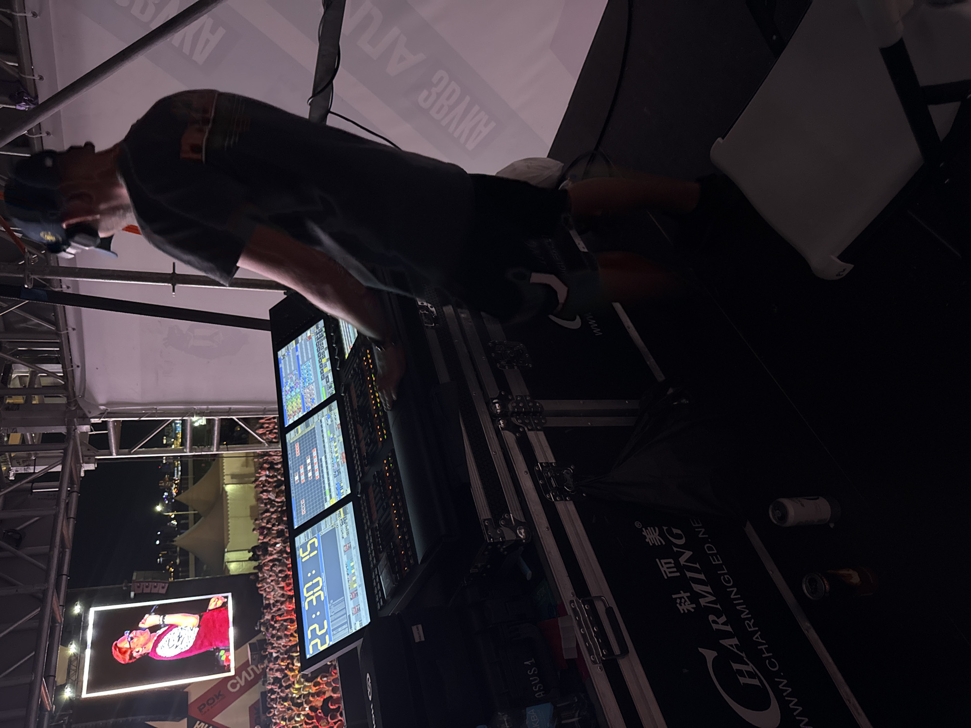 An outdoor nighttime concert is underway, with a large crowd of people visible in the background. On a stage, a person wearing a dark t-shirt, black shorts, and a dark baseball cap is operating a soundboard. The soundboard has multiple screens displaying data and controls, including a prominent timer showing "22:30:15". Stage lighting illuminates the scene, casting shadows and highlighting the equipment. In the distance, tents and city lights are visible, suggesting an urban or suburban setting. This event is taking place in Khabarovsk, Russia.
