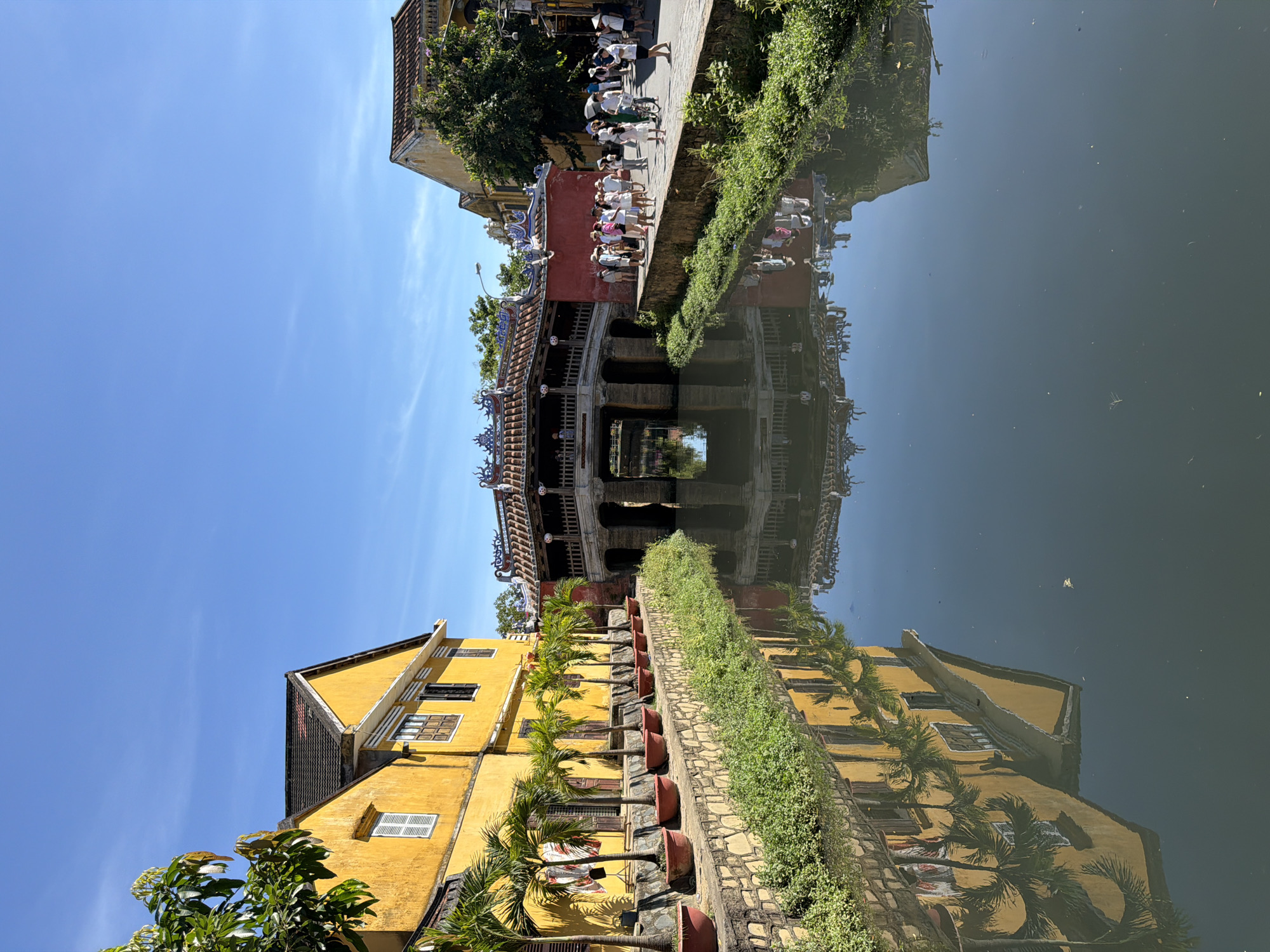 The image depicts a scenic canal in Hoi An, Vietnam, reflecting the architecture and vegetation. On the left, a bright yellow building with a dark tiled roof and white-shuttered windows lines the canal. Several potted palm trees are arranged along the building's stone facade.
Dominating the center is the iconic Japanese Covered Bridge, a historic structure with a pagoda-like roof and ornate decorations. The bridge spans the canal, with arched openings allowing water to flow underneath.
On the right side of the canal, a pathway is populated by a group of people, mostly wearing hats, strolling and observing the bridge and surroundings. A bicycle is also visible amongst the pedestrians. Lush green vegetation borders the canal on both sides. The sky is a clear, vibrant blue. The water surface acts as a mirror, vividly reflecting the yellow building, palm trees, the bridge, and the people.