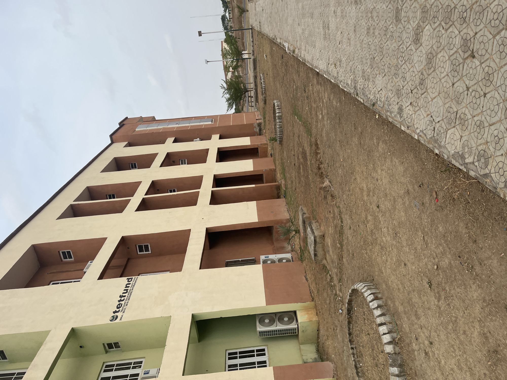 The image shows the exterior of a multi-story building in Baganakwo, Nigeria. The building has a light yellow facade with brown accents on the recessed balconies. The "tetfund" logo is visible on the building's side. Several air conditioning units are mounted on the exterior walls. The building is adjacent to a dirt area with stone borders, and a paved walkway with a patterned surface runs along the edge of the dirt area. In the background, there are more distant buildings and streetlights under an overcast sky. No people are visible in the image.