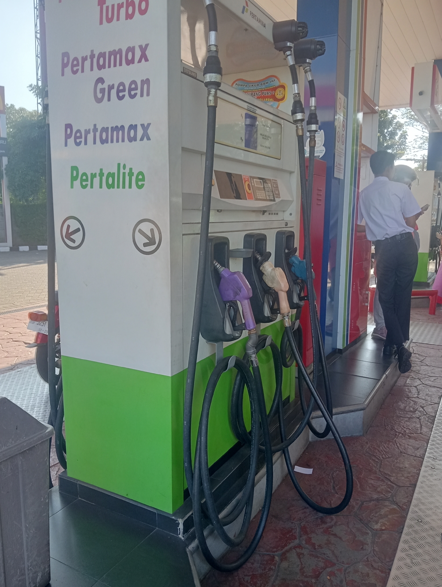 The image captures a Pertamina gas station in Surabaya, Indonesia, showcasing multiple fuel dispensers. The primary focus is on a white fuel pump with various fuel types labeled: "Turbo," "Pertamax Green," "Pertamax," and "Pertolite." Four fuel hoses with different colored nozzles hang from the pump, with a purple, a light brown, and a blue nozzle visible. To the right of the pump, a person wearing a white shirt and black pants is seen, facing away from the camera, possibly a customer or an attendant. The ground is paved with textured reddish-brown tiles, and a portion of a motorcycle with a license plate reading "L 3251 12-23" is visible on the left. A gray trash bin is also present. The overall scene depicts a typical transaction point at a gas station.
