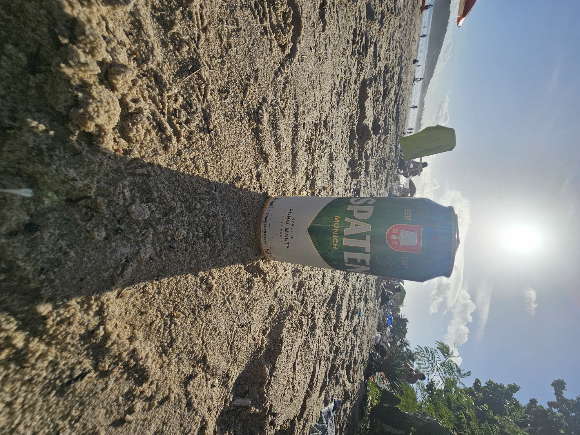 Here is a description of the image:
The image shows a can of Spaten beer on a beach. The can is lying on its side in the sand, with its label facing up. The beach appears to be crowded in the background, with several people and a few umbrellas in the distance, along the shoreline. The sky is a bright blue, and the sun is high in the sky. Trees and other vegetation are visible at the edge of the beach.
Given the label on the can, the location is likely in Brazil, as the label includes the words "CERVEJA PURO MALTE" written in Portuguese. While the city is not stated on the can, "Armacao dos Buzios, Brazil" has been provided in the prompt.