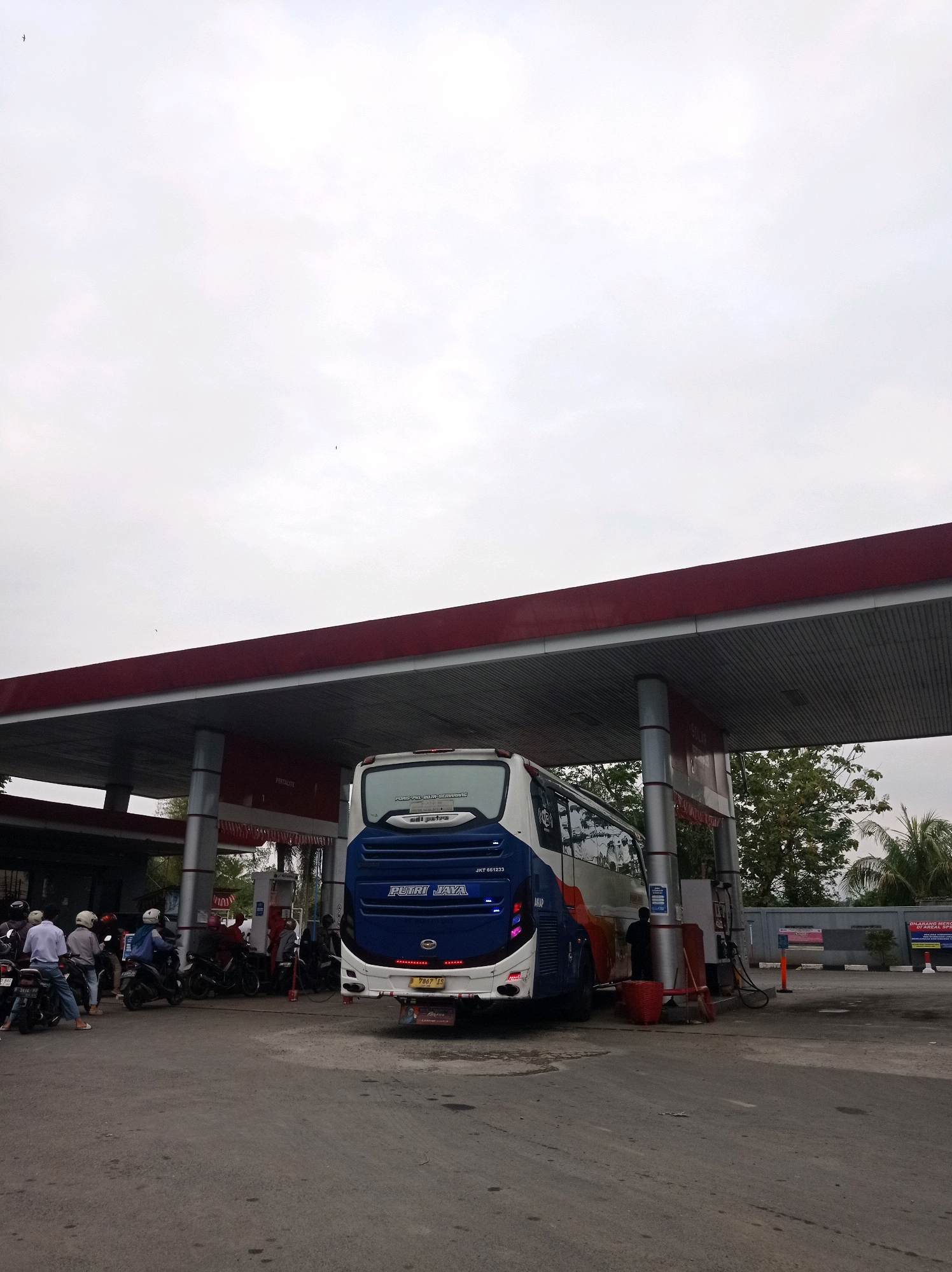 The image shows a bus and several motorcycles at a gas station in Bojong, Indonesia. The bus, a white and blue "PUTRI JAYA" coach, is parked at a fuel pump. Several people wearing helmets are on or near their motorcycles, appearing to be waiting or refueling. The gas station has a red roof supported by grey pillars, with visible signage indicating "PERTALITE". Trees and a cloudy sky form the background. The ground is asphalt, showing signs of wear. A red basket and a broom are visible near the bus. A sign in the background reads "DILARANG MEROKOK DI AREA SPBU".
