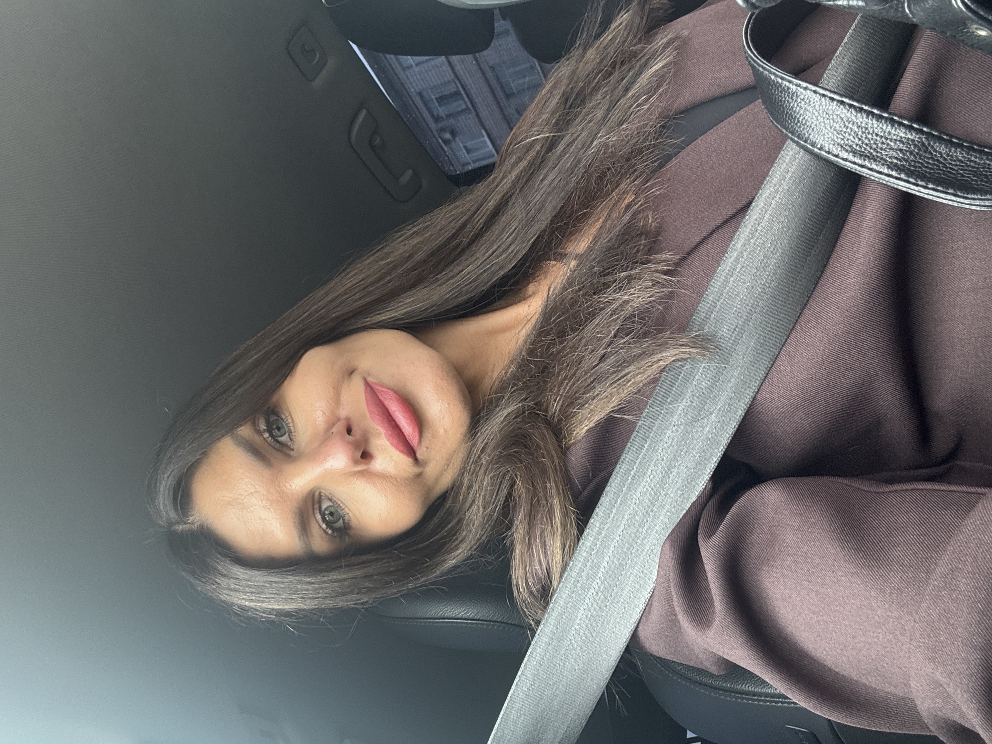 A woman with long, dark brown hair and fair skin is seated in the driver's seat of a car, wearing a dark brown blazer over a black top and a grey seatbelt. She is looking at the camera with a slight smile. Her makeup includes bold lipstick and eyeliner. The car's interior is visible, with the car's ceiling and a portion of the windshield. Outside the car, a building is visible through the windshield, suggesting an urban environment. The specific location of Chaikovsky, Russia, cannot be determined from the visual information provided in the image.