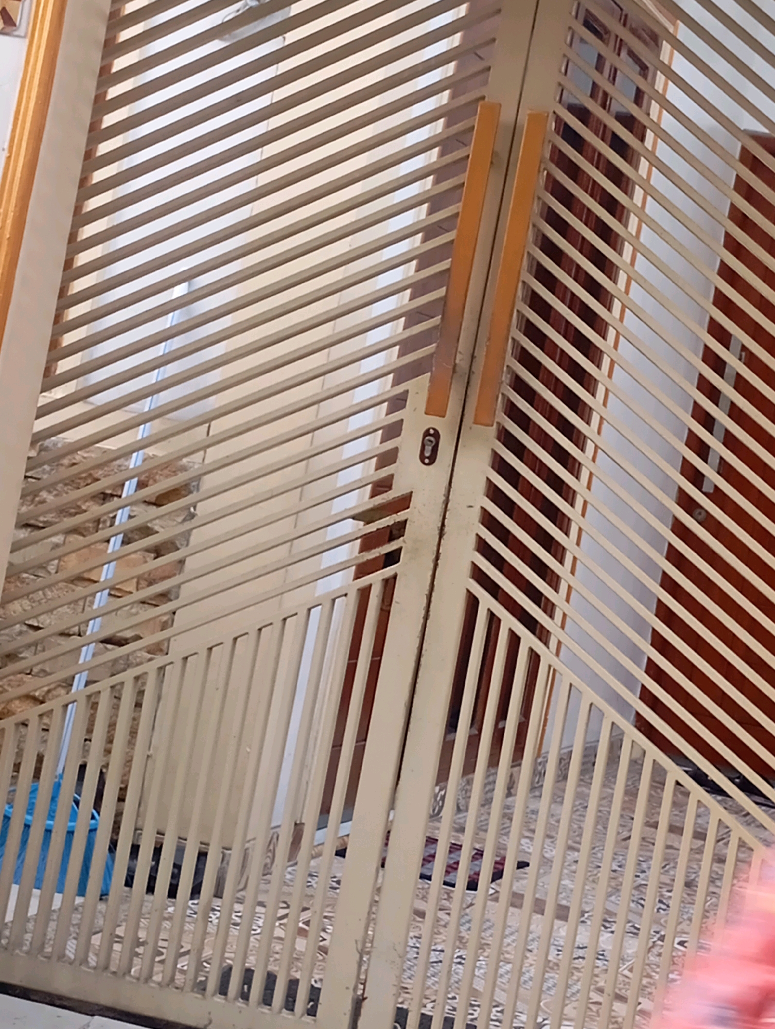 The image shows a closed, light-colored metal gate with diagonal slats. The gate is made of vertical bars and horizontal slats, with two vertical poles on the left side. There appears to be a keyhole or latch mechanism on the left pole. Behind the gate, a stone wall is visible on the left, and a darker, paneled surface, possibly a door or wall, is visible on the right. The ground in front of the gate has a patterned surface, and a blue object, possibly a trash bag, is visible near the bottom left. The image is taken from a slightly low angle, looking up towards the gate. This scene is located in Babelan, Indonesia.