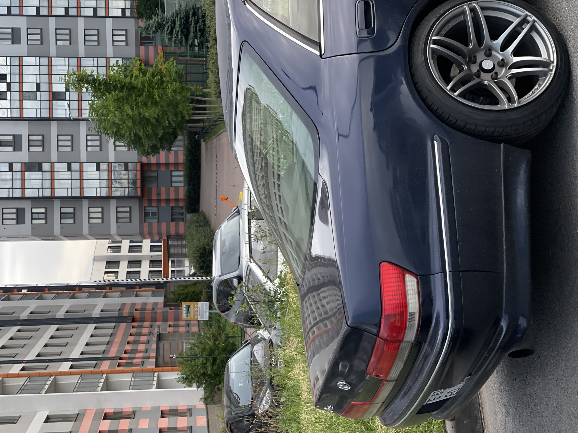 The image shows the side and rear of a dark blue sedan parked on a paved surface next to a grassy area. The car has a silver alloy wheel visible on the right side. In the background, there are several modern apartment buildings with many windows. The buildings have a repeating pattern of light grey and orange stripes. Green trees and shrubs are also visible between the buildings and the car. A yellow sign with text and graphics is posted on a pole to the left of the buildings, and a portion of what appears to be a white vehicle is visible behind the dark blue sedan. The sky is overcast, suggesting a cloudy day. The image is taken from a low angle, looking upwards towards the buildings. There is no visible text that can be clearly read.