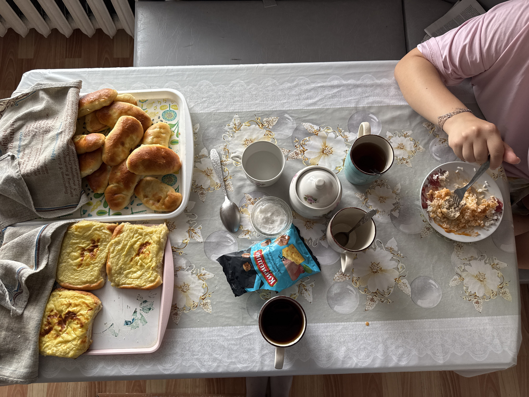 A high-angle, full shot shows a breakfast spread on a table covered with a light-colored tablecloth featuring a floral pattern and lace trim. The table is set with various food items and beverages. To the left, a white tray holds several golden-brown baked goods, possibly small loaves or rolls, covered partially by a beige cloth. Next to it, a pink tray contains three rectangular baked items with a golden-brown, slightly crispy top, appearing to be some form of casserole or baked egg dish.
In the center of the table, a blue and white patterned packet of crackers lies open, next to a small white bowl filled with white granular material, likely sugar or salt. A silver spoon rests beside the bowl. Several cups are visible: a white cup containing dark liquid, possibly coffee or tea, is positioned in the foreground. Another blue and white mug, also with dark liquid, sits further back. A white cup with a floral design, containing a small spoon, is to the left of the sugar bowl. To the right, a white plate holds a portion of rice with some orange sauce and a fork.
A person, wearing a pink t-shirt, is partially visible on the right side of the image, their right arm extended towards the plate of rice. They are wearing a silver bracelet on their wrist.
The background reveals a gray sofa and a radiator. The lighting suggests it is daytime, with natural light casting shadows. The overall scene depicts a domestic breakfast setting. There is no indication of the city of Бородулино, Russia in the image itself.