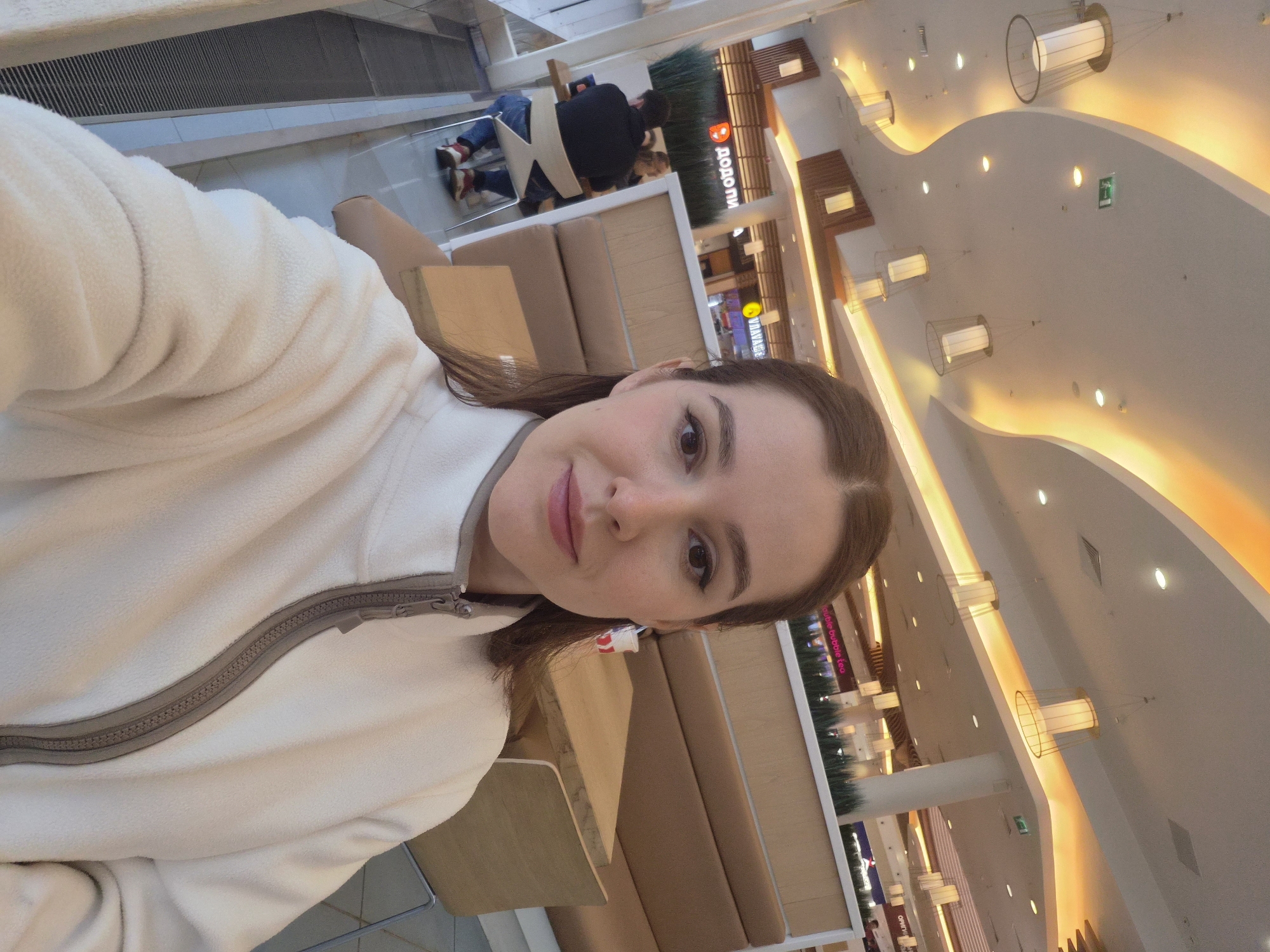 The image shows a young woman taking a selfie in what appears to be a food court area of a shopping mall. The woman is wearing a light-colored fleece jacket. In the background, there are dining tables and booths, as well as various food establishments with signs in Russian, including "ДОДО ПИ" and "UNAVAGA." There are also people seated at tables in the background. The ceiling features modern lighting fixtures and architectural design elements, suggesting an indoor commercial space. The overall environment appears to be clean and well-lit. The location is identified as the city of Saint Petersburg, Russia.