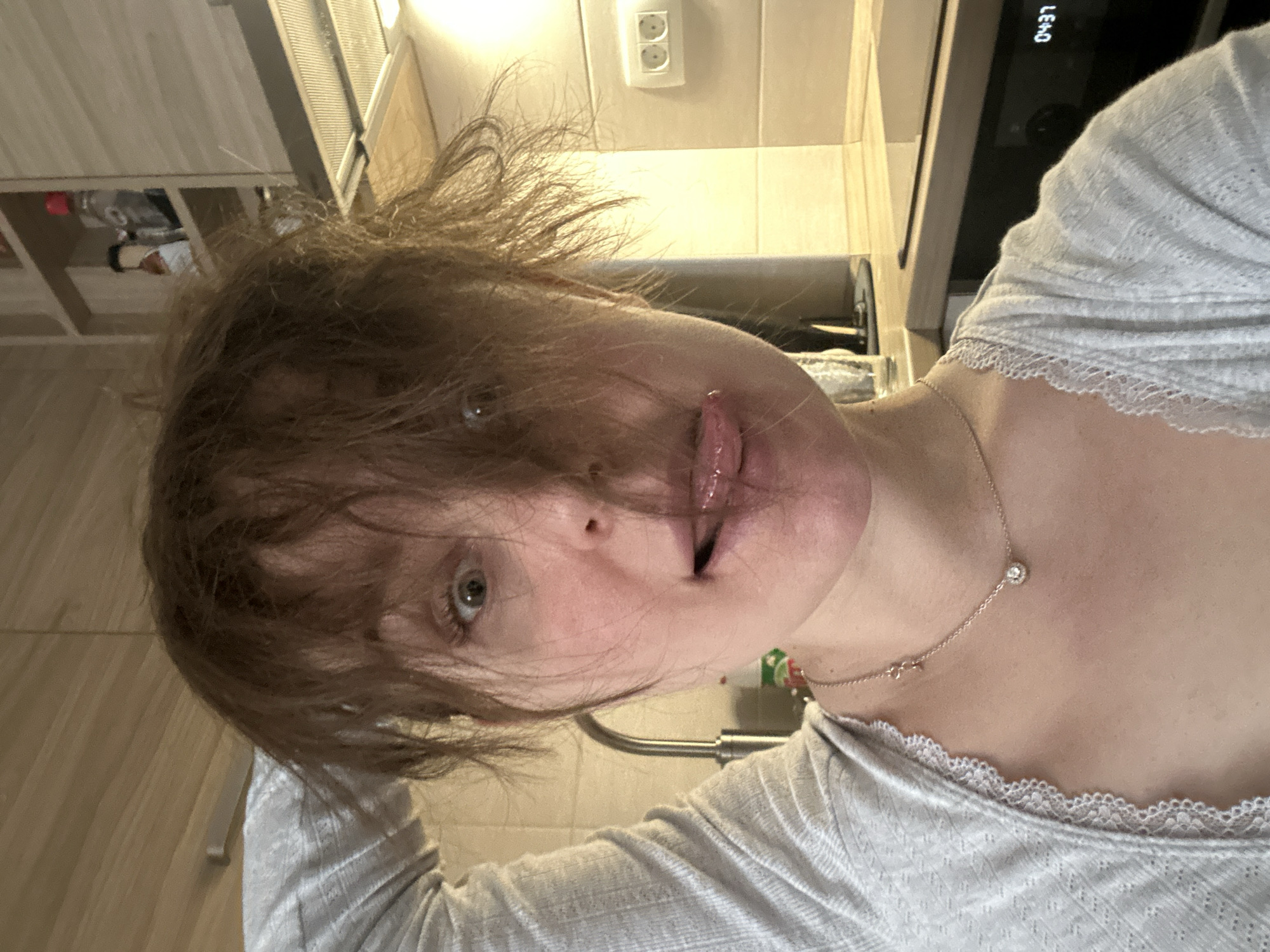 A close-up image depicts a female individual positioned in a residential kitchen in Budva, Montenegro. The subject has disheveled reddish-brown hair, with strands partially covering her forehead and left eye. Her right eye is wide open, and her mouth is open with her tongue extended towards her upper lip. She wears a light grey, ribbed top with white lace trim along the neckline and a gold-colored necklace featuring a small circular pendant. Her left arm is raised, with the forearm and hand obscured. The background reveals a kitchen area with light wood-grain wall cabinets and countertops, light-colored tiled walls, and a stainless steel sink faucet. A bottle with a green and white label is partially visible near the sink. An overhead range hood with a metal filter is present. To the right, a white double electrical outlet is mounted on the tiled wall, and below it, a dark-colored appliance, likely an oven, displays a digital readout of "04:37".