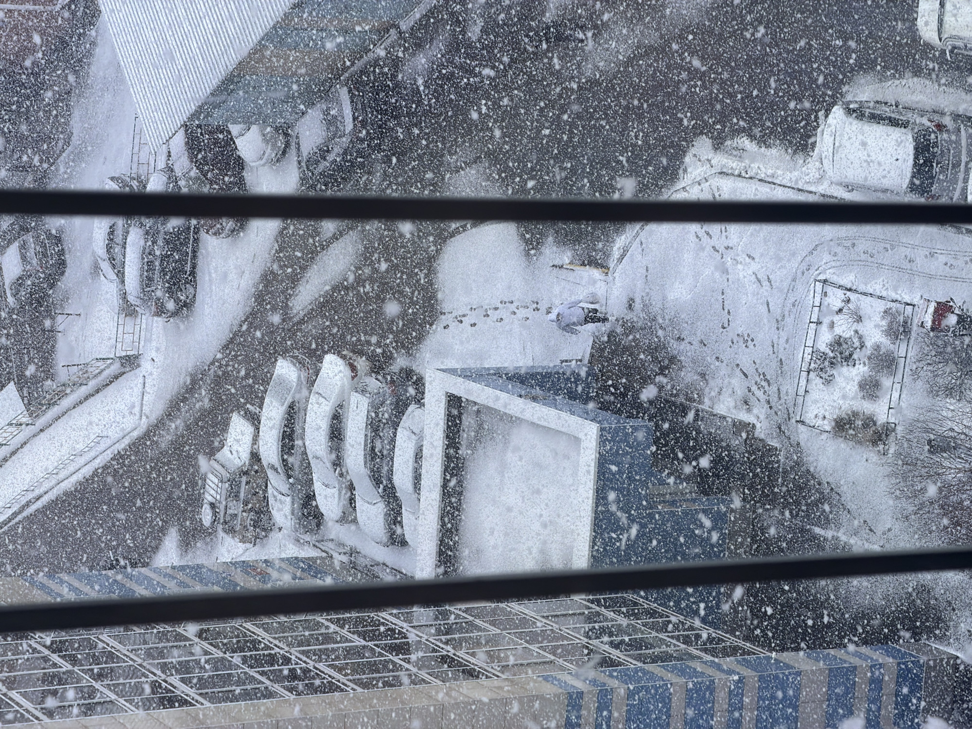 An overhead shot through window blinds shows a snow-covered area in Krasnoyarsk, Russia. The area features parked cars, walkways with footprints in the snow, playground equipment, and a modern art installation of white abstract shapes. The blizzard is intense.