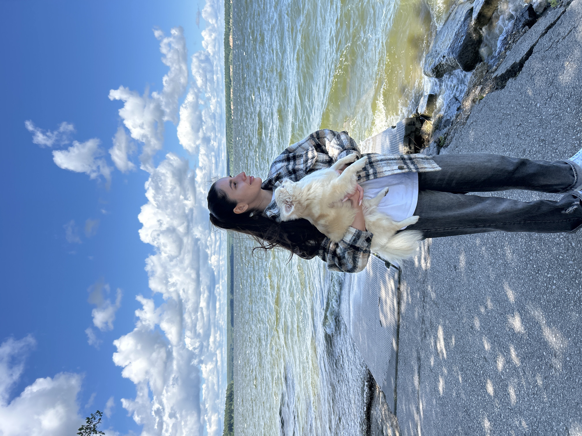 A young woman with dark hair pulled back in a ponytail stands outdoors, holding a small, light-colored dog. She is wearing a plaid jacket over a white t-shirt and dark jeans. The woman is looking to her right, with a slight smile. The dog is facing towards her.
They are positioned on a paved walkway next to a body of water. The walkway is grey and has a diamond plate pattern in some sections. To the right of the walkway, there are large rocks and the choppy, green water of a lake or river. The water is reflecting the bright blue sky and white, puffy clouds. In the distance, a tree line is visible on the opposite shore.
The lighting suggests it is daytime, with dappled sunlight casting shadows on the walkway. The overall scene is bright and outdoors. The location is identified as the embankment of the Tom River in Mezhdurechensk, Russia.