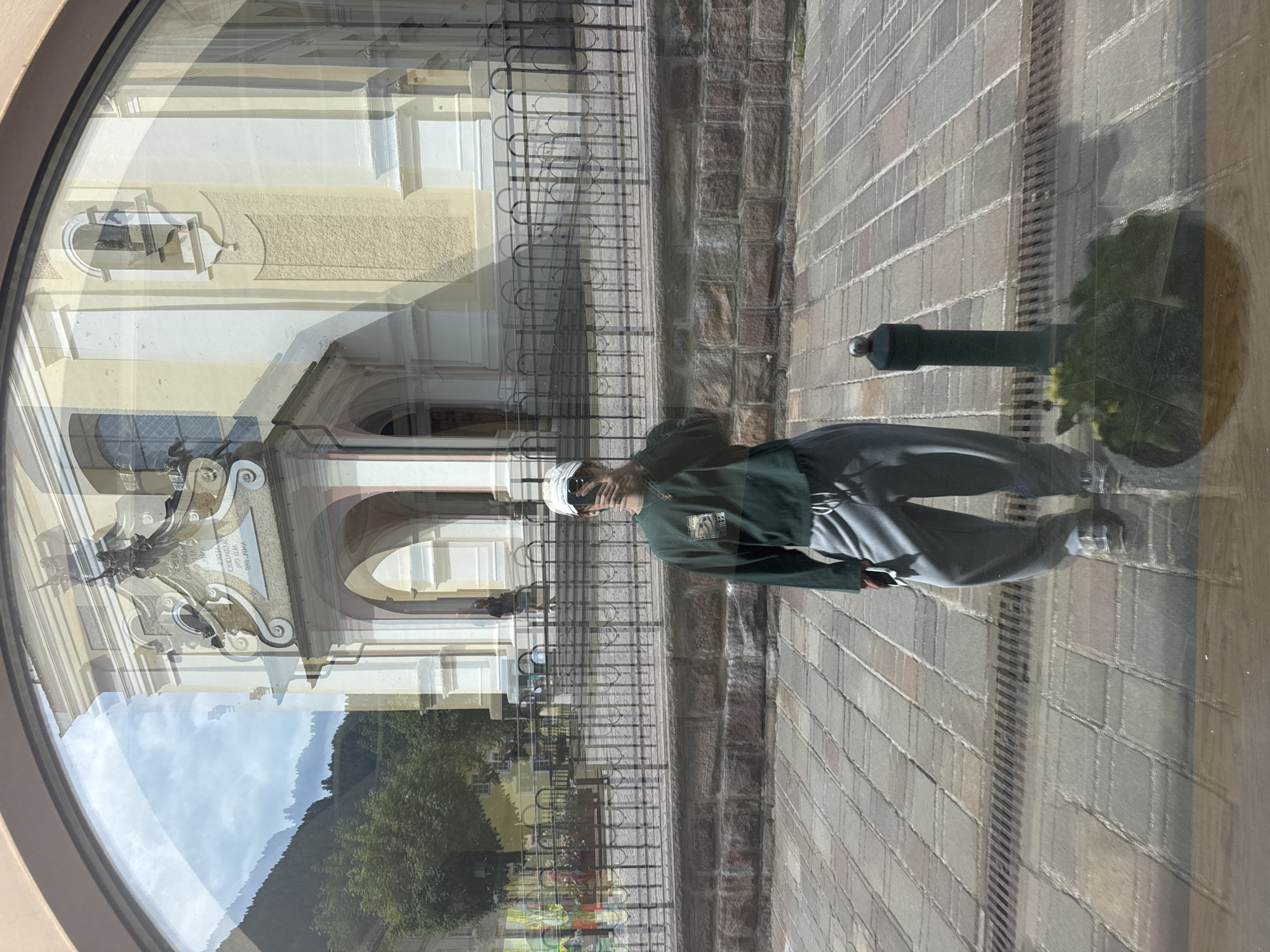 The image is a reflection in a curved window, showing a person standing in front of a building and a stone-paved area. The person is wearing a dark green jacket, grey pants, and a white cap. They are looking towards the camera. In the reflection, the building appears to be made of light-colored stone with arched entrances and ornate architectural details. A black metal fence is visible in front of the building. The stone-paved area is made up of rectangular bricks. The time of day appears to be daytime, with bright lighting and shadows suggesting a sunny day. The reflection also shows a glimpse of a mountainous landscape with green trees and a cloudy sky, indicating a natural setting. The overall mood is casual and perhaps a bit artistic due to the reflective surface. There are no discernible words or text in the visible portion of the image.