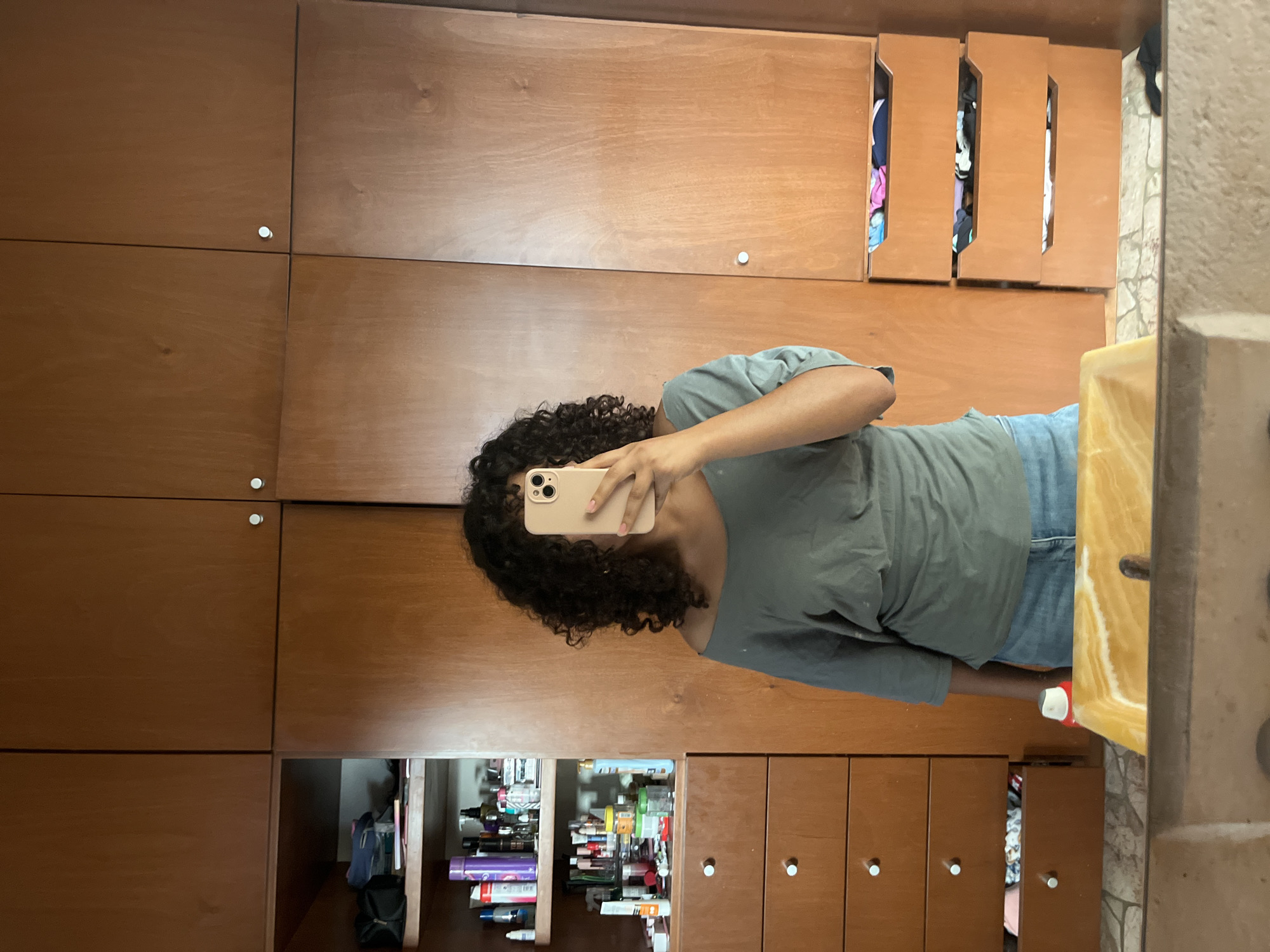 A person with curly hair is taking a selfie in front of a large wooden cabinet with multiple doors and drawers. Some doors are open revealing contents such as clothes and cosmetic products. The location is Unknown, Unknown. The person is wearing a grey t-shirt and blue jeans. They are holding a beige smartphone to take the photo. A partially visible bathroom sink is in the right part of the frame.