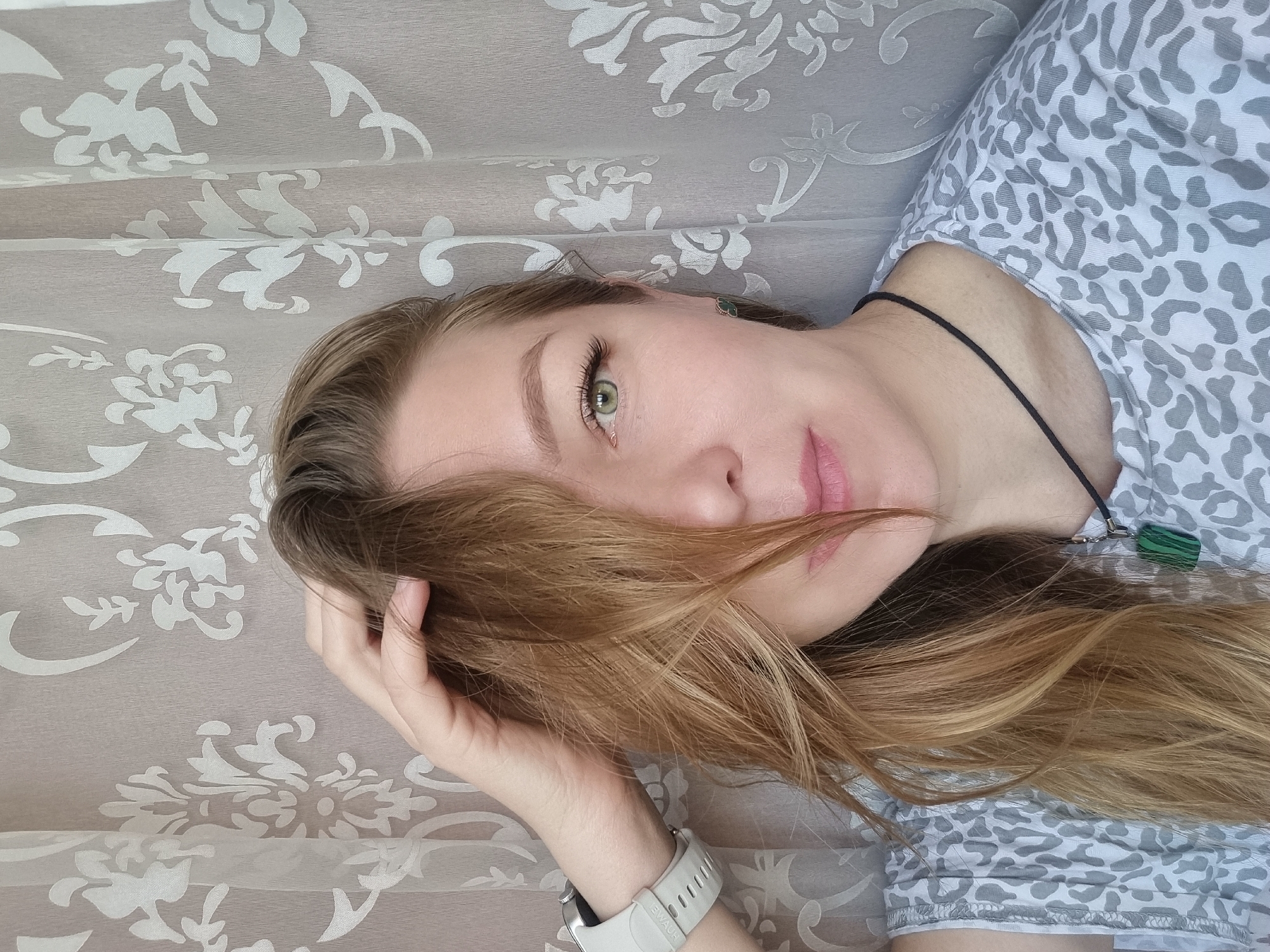 A close-up, portrait-style photograph, rotated 90 degrees clockwise, depicts a person, likely a woman, lying down and looking towards the camera. The setting appears to be indoors in Chaikovsky, Russia.
The subject has long, wavy light brown hair, which partially covers the right side of her face. She has prominent light green eyes and light-colored skin. Her expression is neutral and calm, with a slight hint of a smile. Her left hand is raised, with fingers gently touching her hair near her head.
She is wearing a light-colored top with a grey and white leopard print pattern. A black cord necklace with a dark green, rectangular pendant is visible around her neck. In her visible right ear, there is a small dark green stud earring. On her left wrist, she wears a white smartwatch with a white band, on which the text "SWAUH" is visible.
The background consists of a light grey or beige fabric, possibly curtains or a textured wall covering, featuring a recurring white damask or floral pattern. The lighting suggests it is daytime, with soft, natural light illuminating the scene.