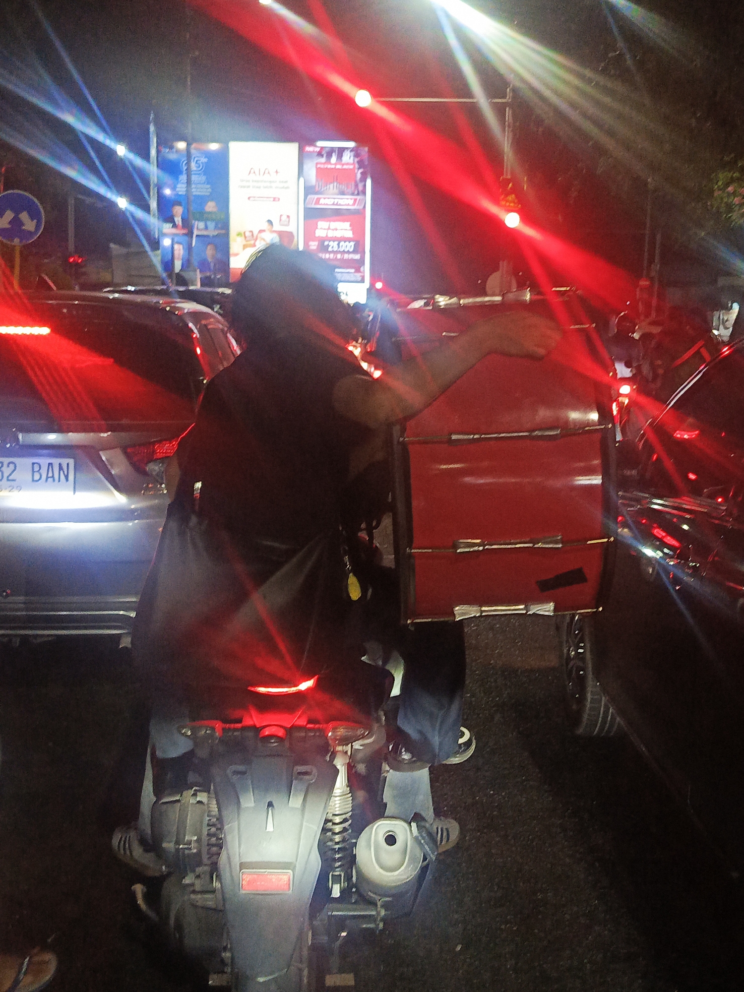 The image shows a person on a motorcycle at night, stopped in traffic. The person is wearing a dark shirt and blue jeans. They have a large red insulated delivery box on the back of the motorcycle. The motorcycle is silver and black. There are cars in front of and behind the motorcycle. The background is a street with billboards and lights. The scene is likely in an urban area.
