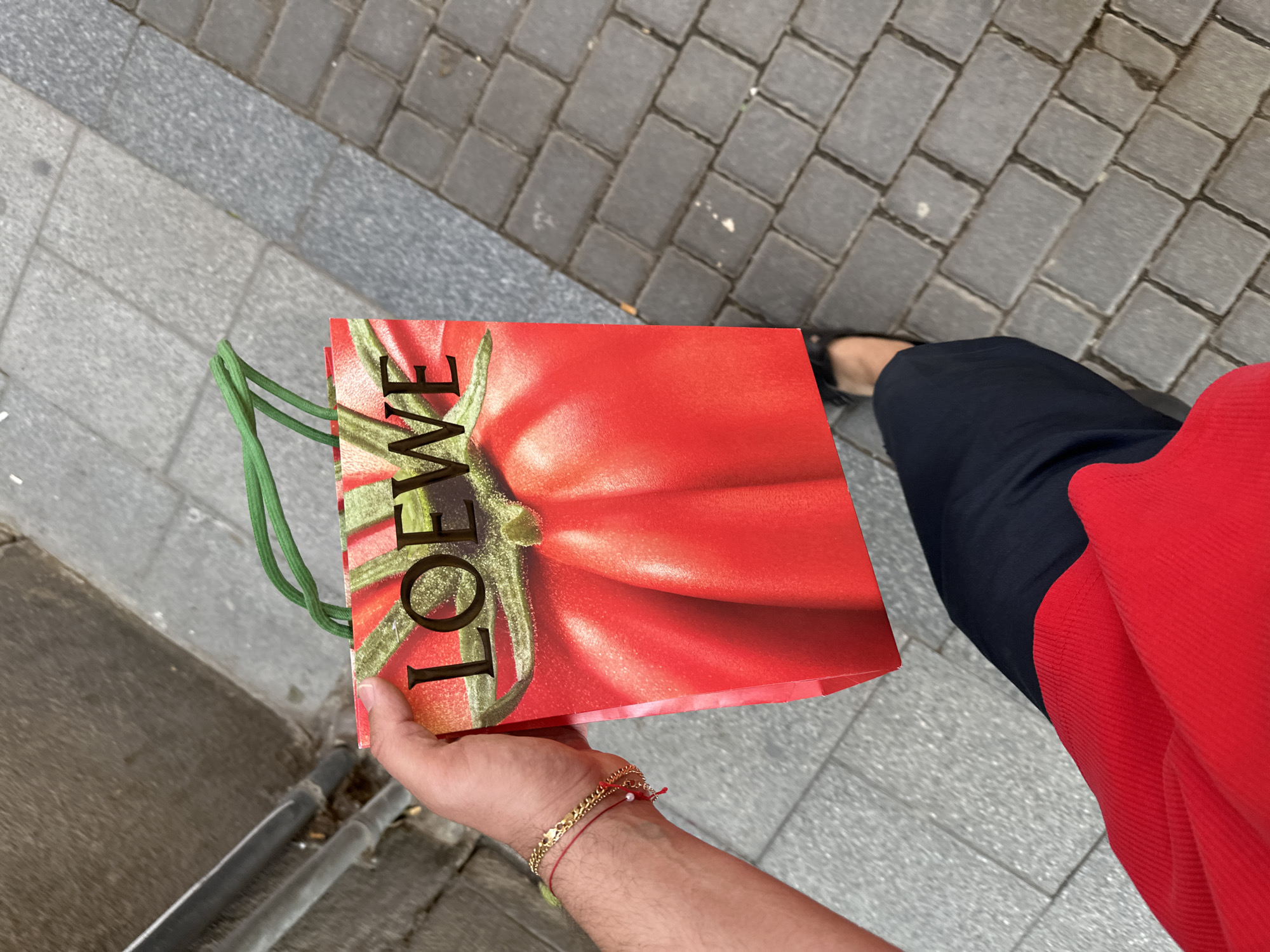 Here is a description of the image:
A person is holding a Loewe shopping bag with a large tomato print on it while walking on a sidewalk. The bag has green handles. The person is wearing a red shirt and navy blue pants. Their hand is visible, adorned with a gold bracelet and a red string bracelet. The sidewalk is made of gray interlocking paving stones. The image appears to be taken from above, looking down at the person and the bag.
