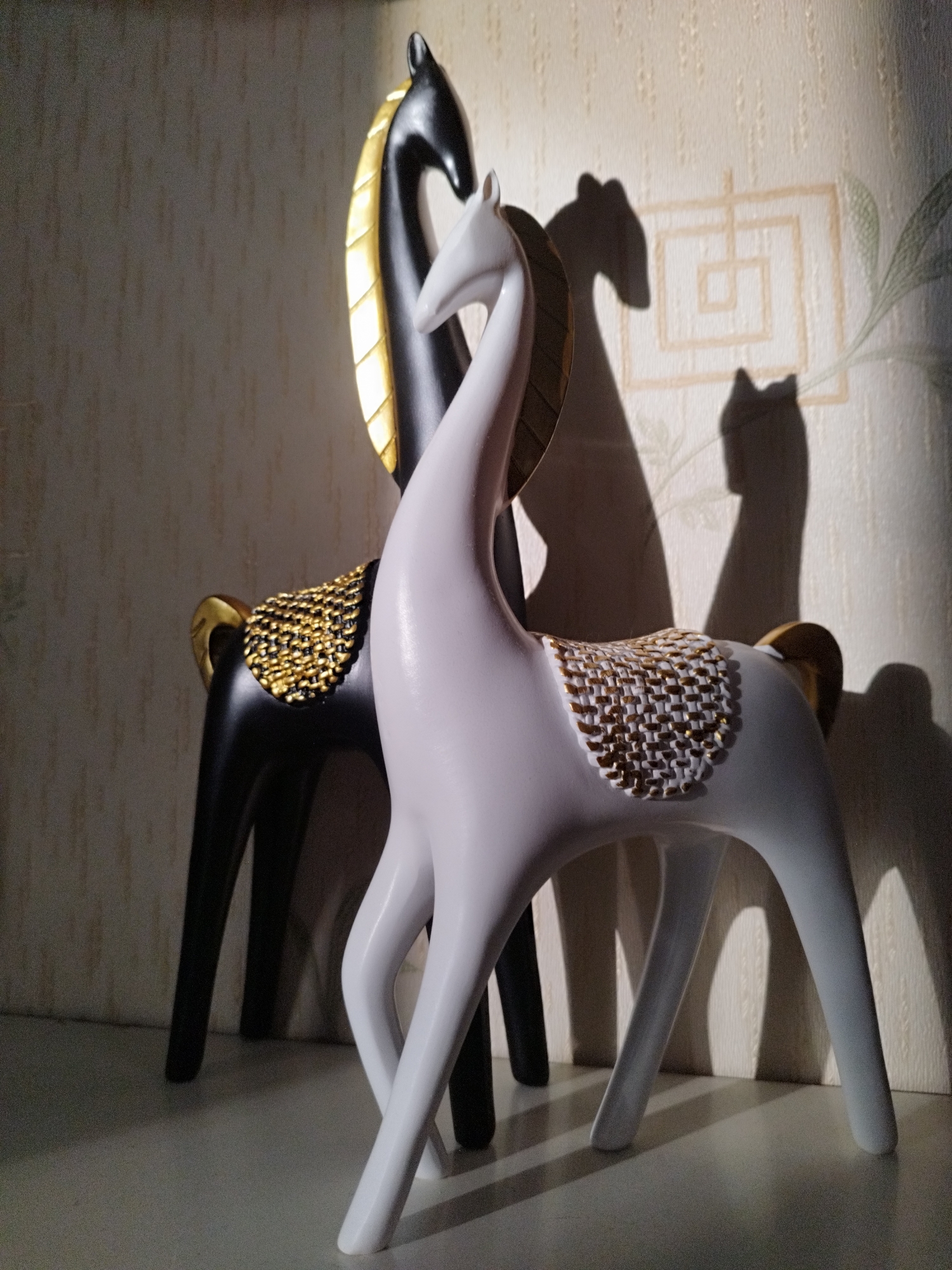 Two decorative horse figurines stand on a white surface against a textured wallpapered wall. The figurine in the foreground is white with gold accents on its back, resembling a saddle. The figurine behind it is black with gold accents on its mane and saddle. Both figurines are illuminated from the side, casting long shadows on the wall behind them. The wallpaper has a repeating pattern and some faint leaf-like shadows. The lighting creates a dramatic effect, highlighting the contours of the horses and their shadows. The scene is static and appears to be an indoor setting, possibly a decorative display. No specific location in Saint Petersburg, Russia, can be determined from the image.