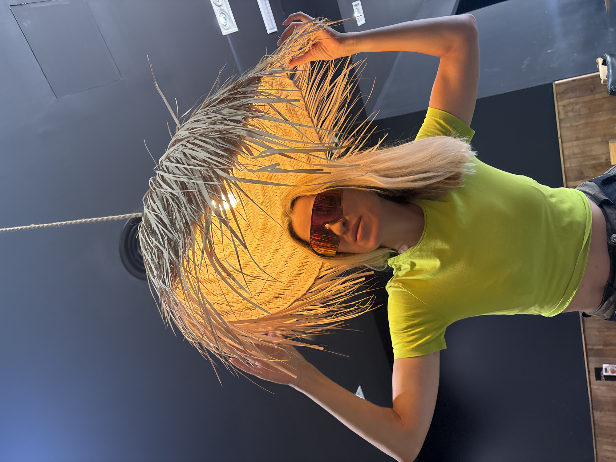 An adult Caucasian female with blonde hair and sunglasses stands indoors, holding up a large, straw-like lampshade over her head with both hands. She is wearing a neon green t-shirt and dark pants. The background is a dark wall with wooden paneling at the bottom. The lighting suggests an indoor setting, possibly a restaurant or shop in València, Spain, given the presence of modern light fixtures on the ceiling. The woman appears to be posing for the photograph.