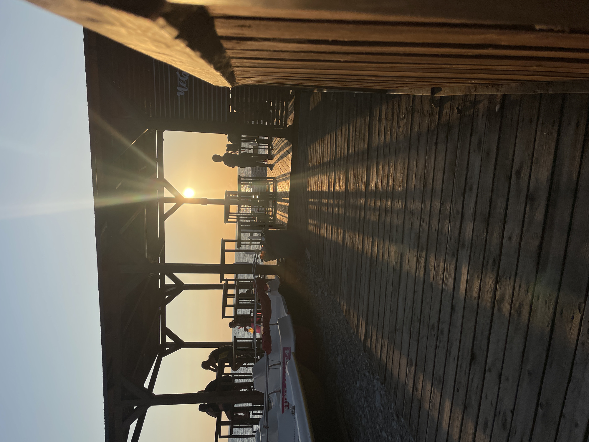 The image depicts a wooden pier by the sea at sunset. Several pedal boats are moored on the left side of the pier. In the background, under a wooden canopy, several people are silhouetted against the setting sun, which is casting long shadows across the pier. One individual stands near the edge of the pier, facing the sea, while others are seated. The sky is clear and the sea is calm. The overall scene conveys a sense of peaceful evening at a coastal location. While the image does not explicitly state its location, the presence of pedal boats and a pier are common features of tourist areas. The text "we go" is visible on a dark structure in the background. Without further information, it is not possible to confirm or deny if this is Sochi, Russia.