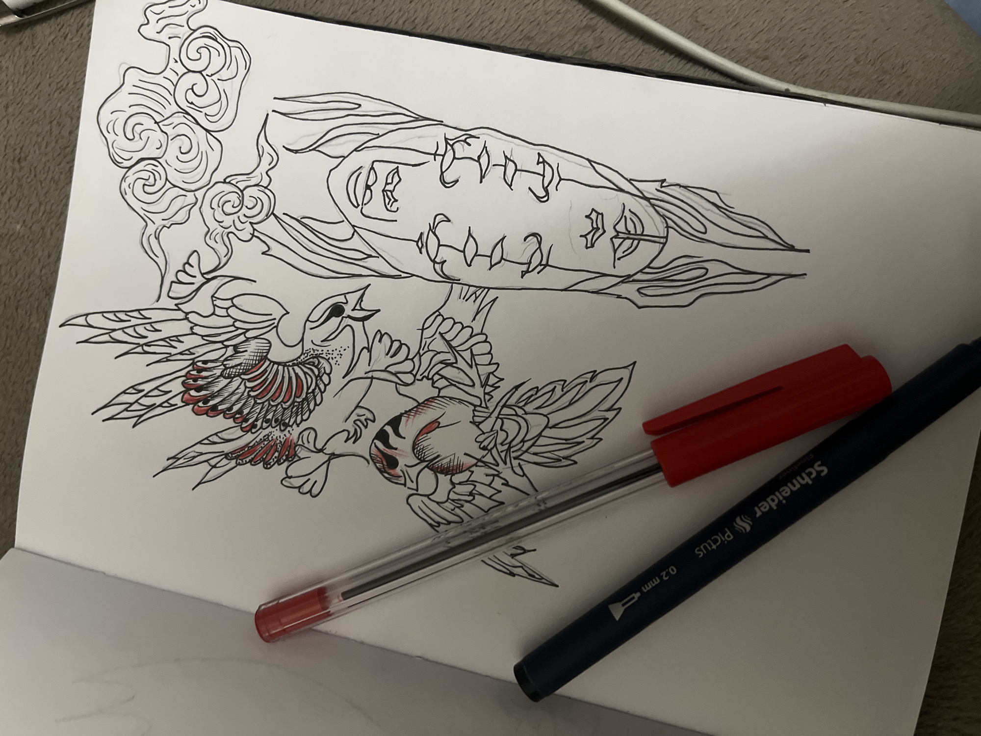 The image shows an open sketchbook on a soft, tan-colored surface. The sketchbook contains a drawing in black ink with red accents. The drawing depicts two birds, possibly jays, in flight. The birds have detailed wing feathers, some highlighted in red. Above the birds is a series of swirling clouds. Above the clouds is a stylized face with several dots. Two pens are lying on the page: a red ballpoint pen with a clear barrel and the cap off, and a black Schneider Pictus 0.2 mm pen. The scene is not located in Sofia, Bulgaria, based on the provided information.