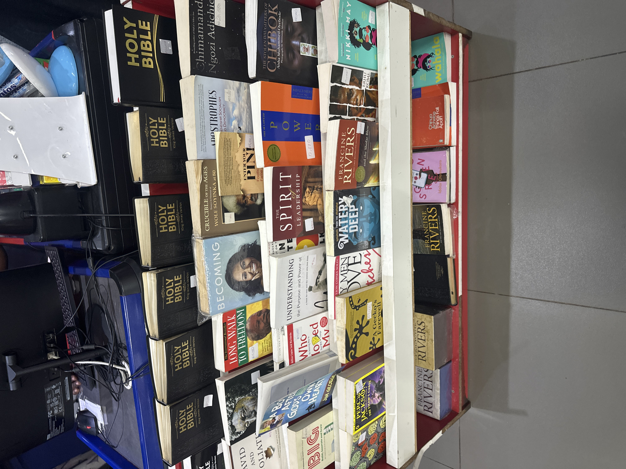 The image shows a bookshelf filled with books in what appears to be a bookstore or a market stall in Barden Yana, Nigeria. The books are arranged in rows on white shelves with red trim. Several books are prominently displayed, including multiple copies of "Holy Bible," "Becoming" by Michelle Obama, "Long Walk to Freedom" by Nelson Mandela, and titles like "The Spirit of Leadership," "The 48 Laws of Power," and "The Stolen Daughters of Chibok." Some books have price tags attached. In the background, a computer monitor, keyboard, and cables are visible, suggesting a functional setup within the establishment. The flooring is tiled with a gray pattern. The lighting appears to be artificial, contributing to a brightly lit indoor environment.