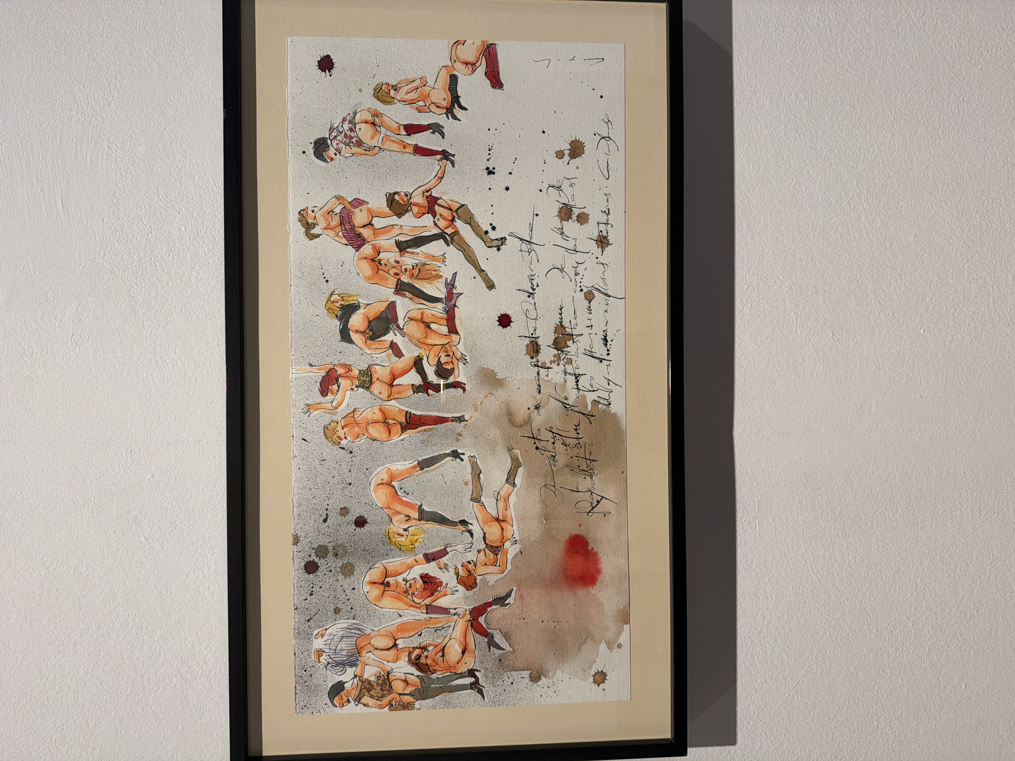 This is a framed artwork depicting a series of figures in a stylized, illustrative manner. The subjects appear to be human figures, predominantly female, depicted in various poses, some of which are acrobatic or contorted. They are rendered with bold outlines and colored with watercolors, giving them a somewhat abstract yet figurative quality. Some figures are nude or semi-nude, wearing lingerie or suggestive clothing. The background of the artwork is a textured paper with scattered ink splatters and watercolor washes, creating a somewhat chaotic or abstract environment.
To the right of the figures, there is handwritten text in a script style, which is difficult to decipher fully. Some words or phrases appear to be present, but their meaning or content cannot be determined from the image alone. The overall impression is that of a dynamic composition, possibly exploring themes of the human form, movement, or sensuality. The artwork is framed with a black border and a cream-colored mat. The setting appears to be an indoor space, possibly a gallery or home, where the artwork is displayed on a textured white wall. The lighting suggests an indoor environment, possibly during the day or with artificial lighting. There are no specific location cues within the artwork itself, other than the stated context of La Cuesta, Spain.